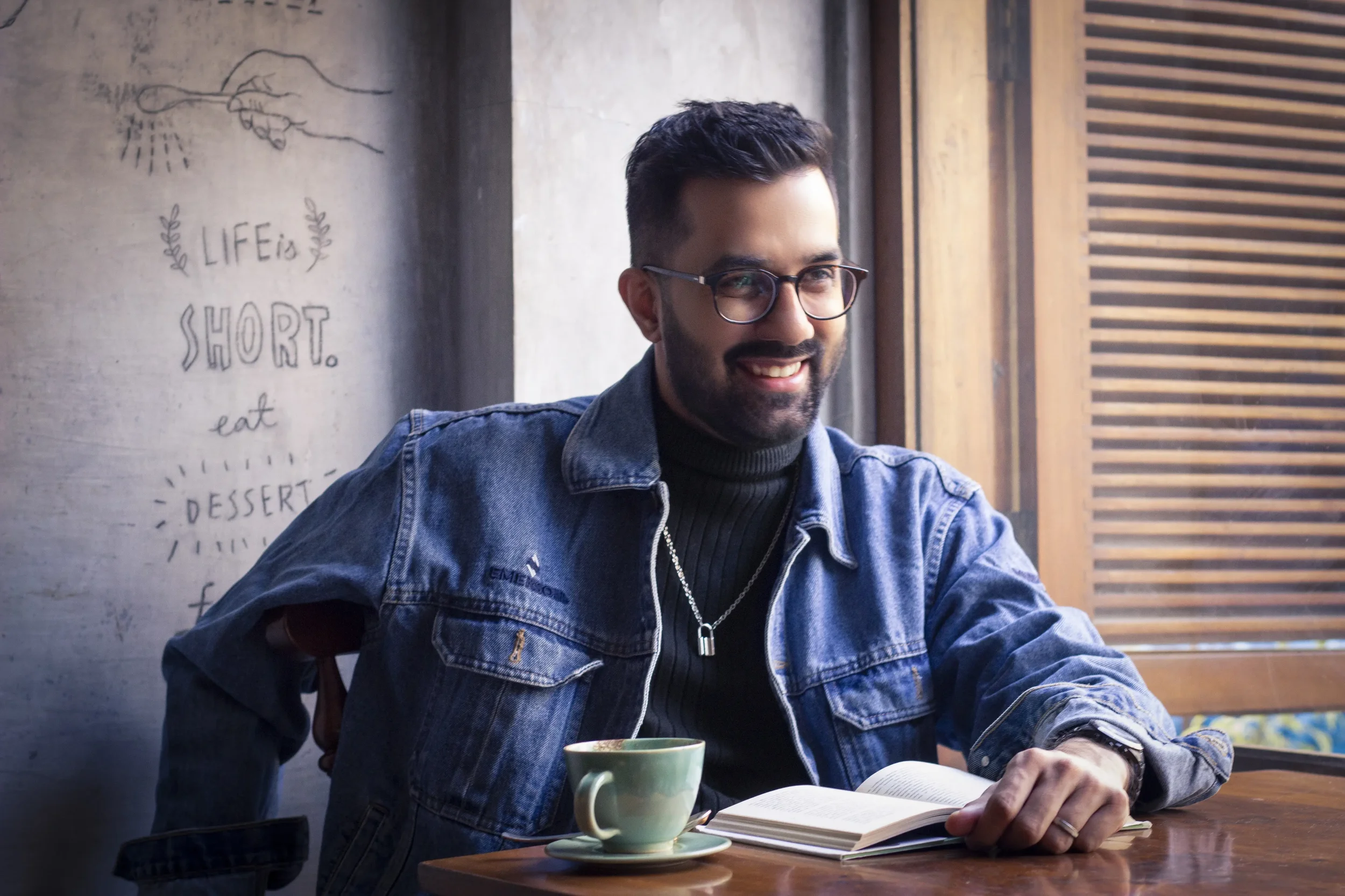 A man with glasses and a beard sitting at a wooden table with a cup and open book, smiling. There is a wall behind him with a hand-drawn art and the words 'Life is short. Eat dessert.'