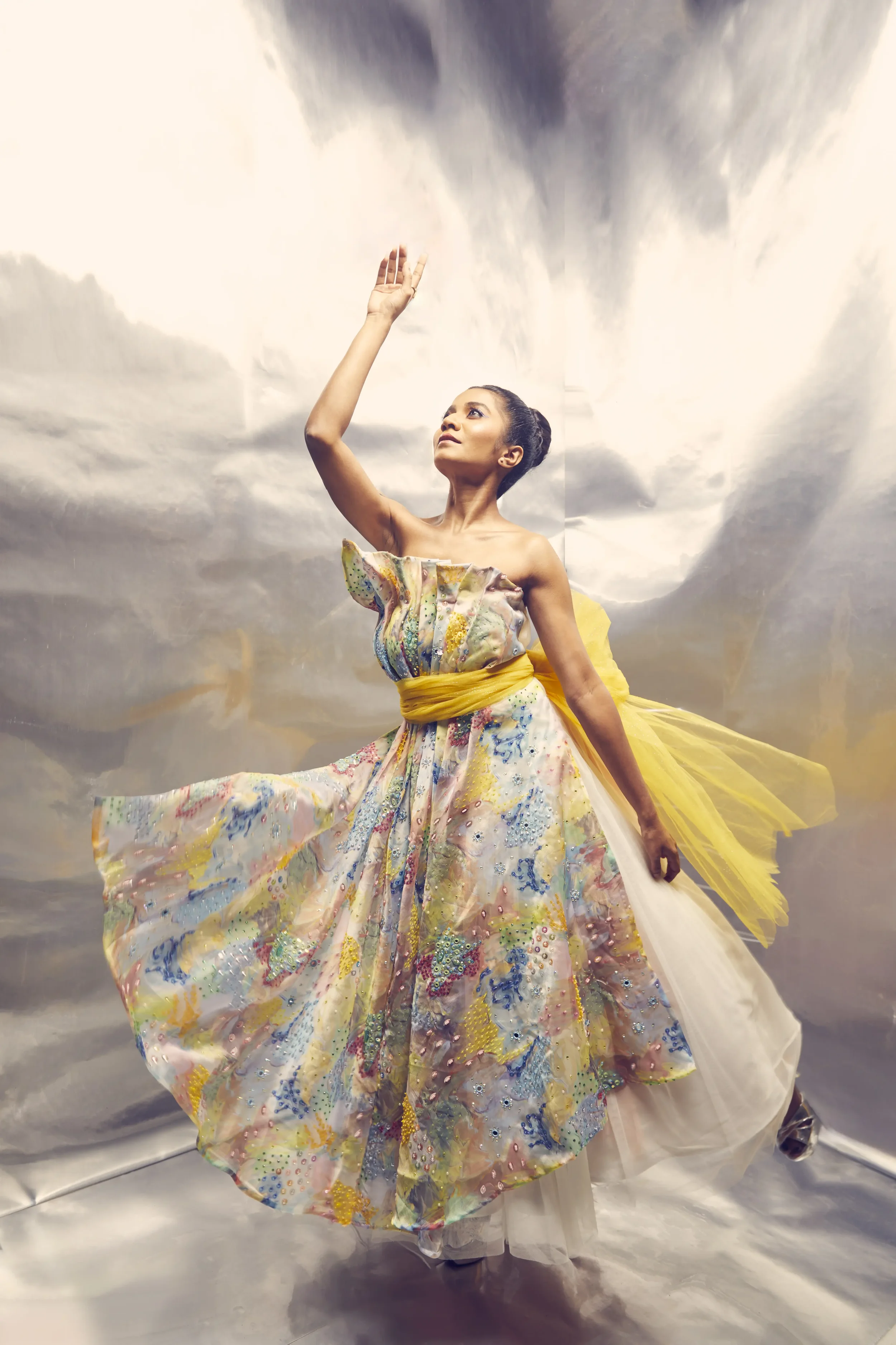 Woman wearing a colorful, patterned dress with a yellow sash, standing against a cloudy sky backdrop, with one arm raised.