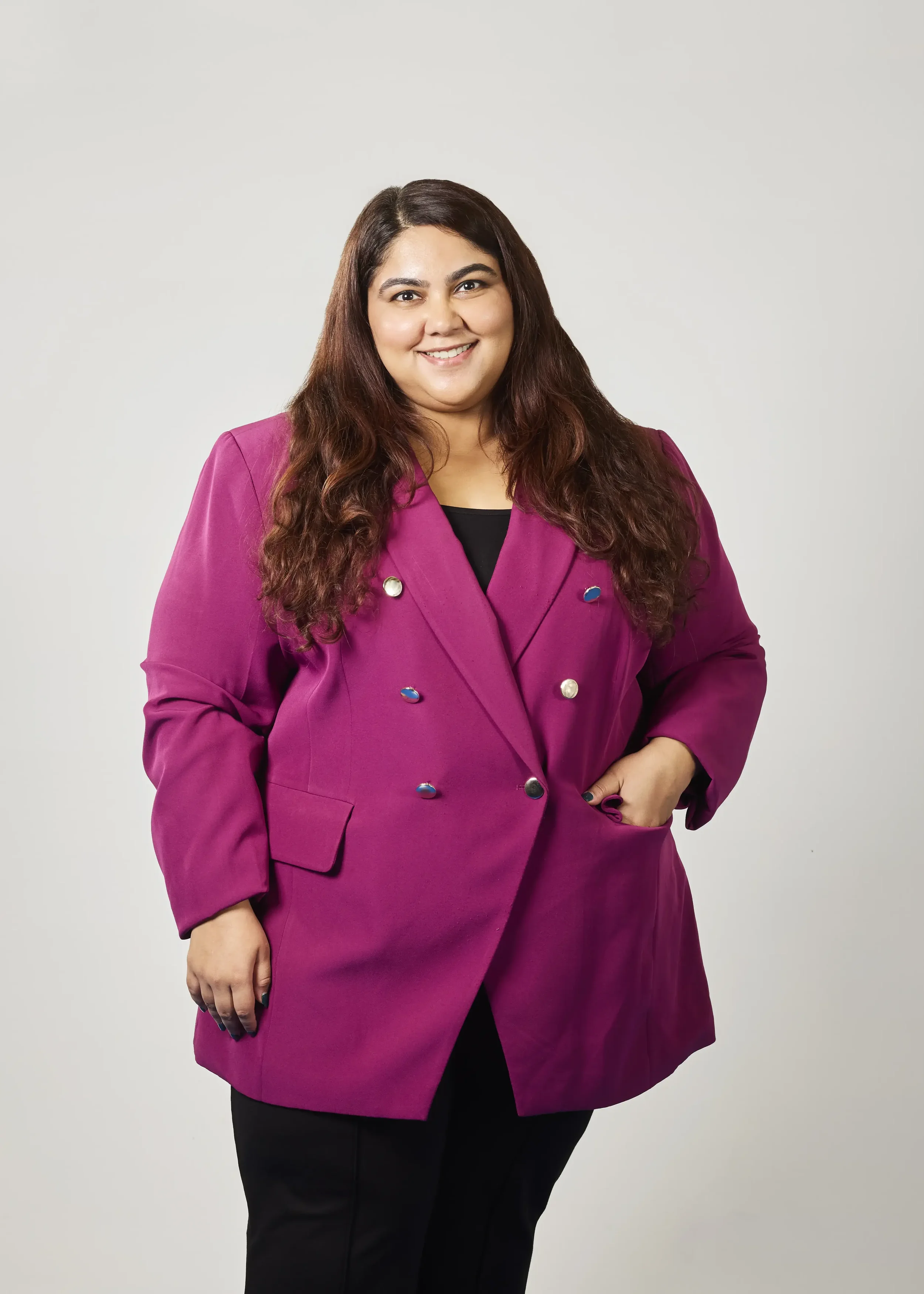 A woman with long, wavy brown hair is smiling and wearing a magenta blazer with decorative buttons over a black top, and black pants, standing against a plain light gray background.