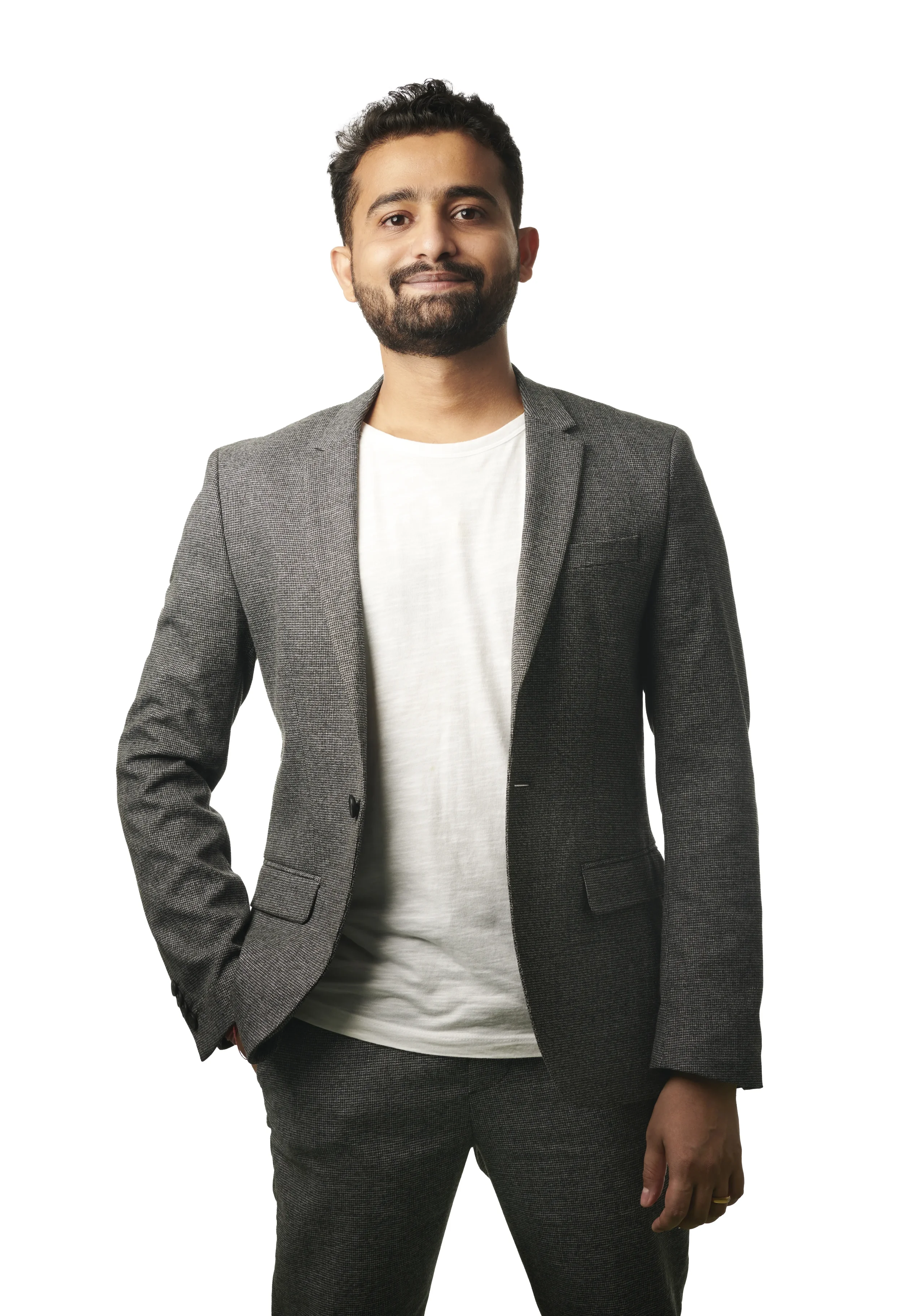 A man with a beard wearing a gray blazer and white T-shirt, standing with one hand in his pocket, smiling against a white background.