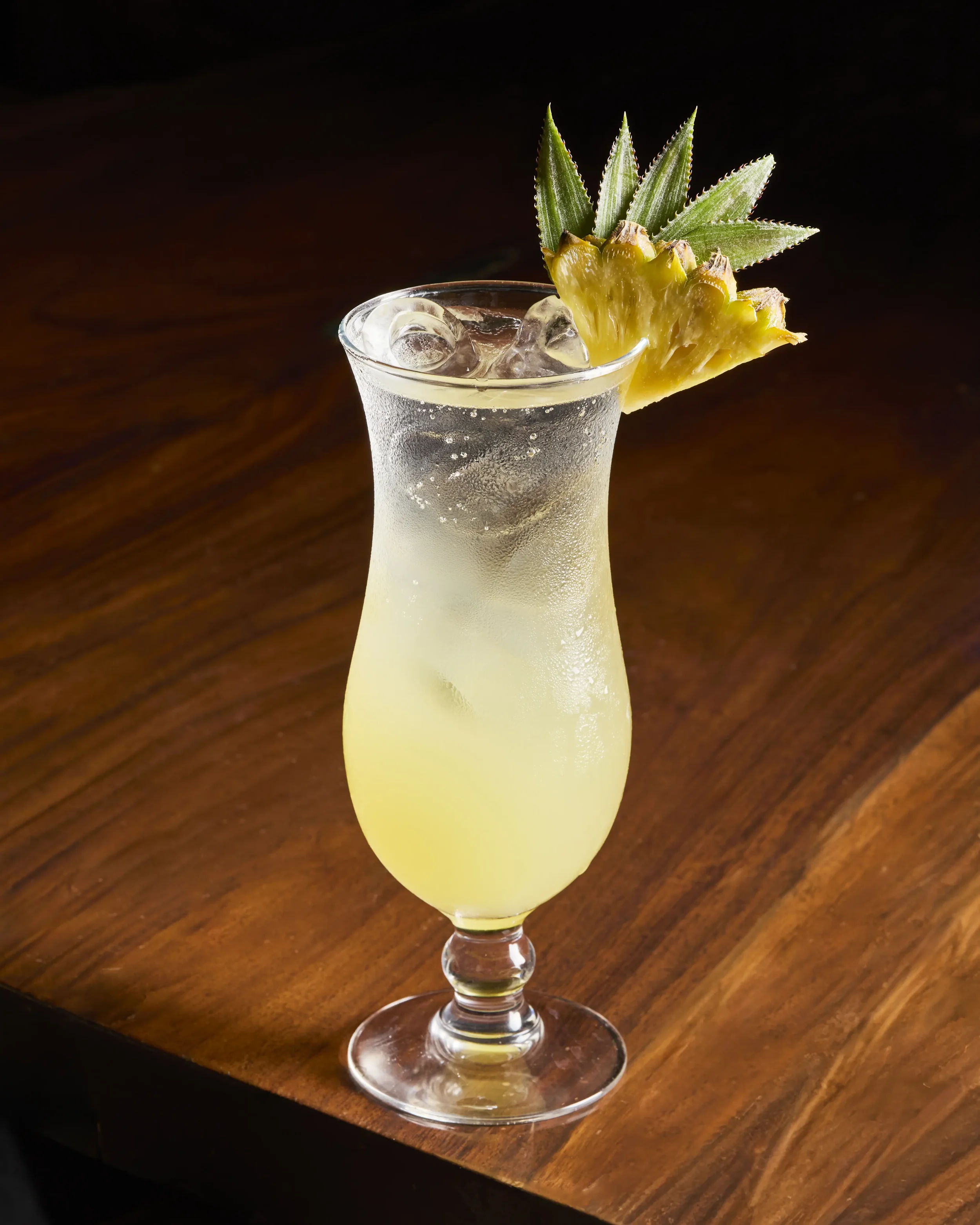 A tropical cocktail in a tall glass with a pineapple wedge and green pineapple leaves garnish, placed on a wooden surface.