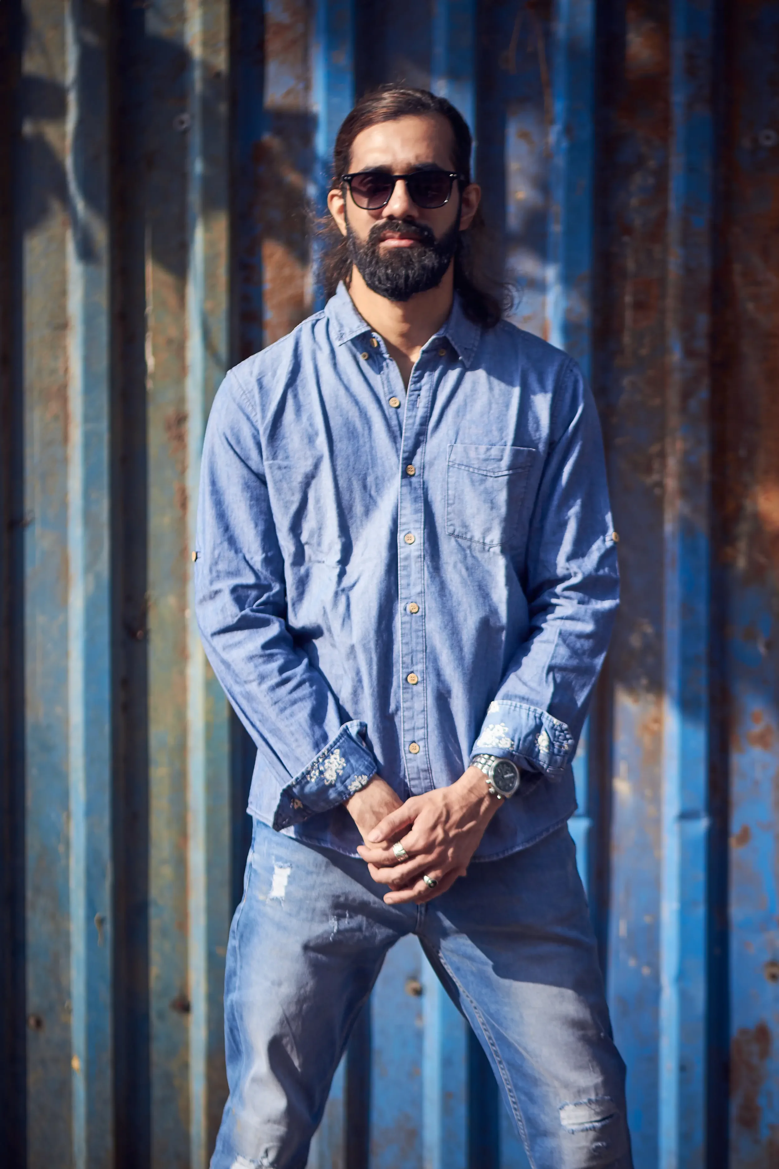 A man with long hair, beard, and sunglasses standing in front of a rusted metal wall, wearing a denim shirt and jeans.