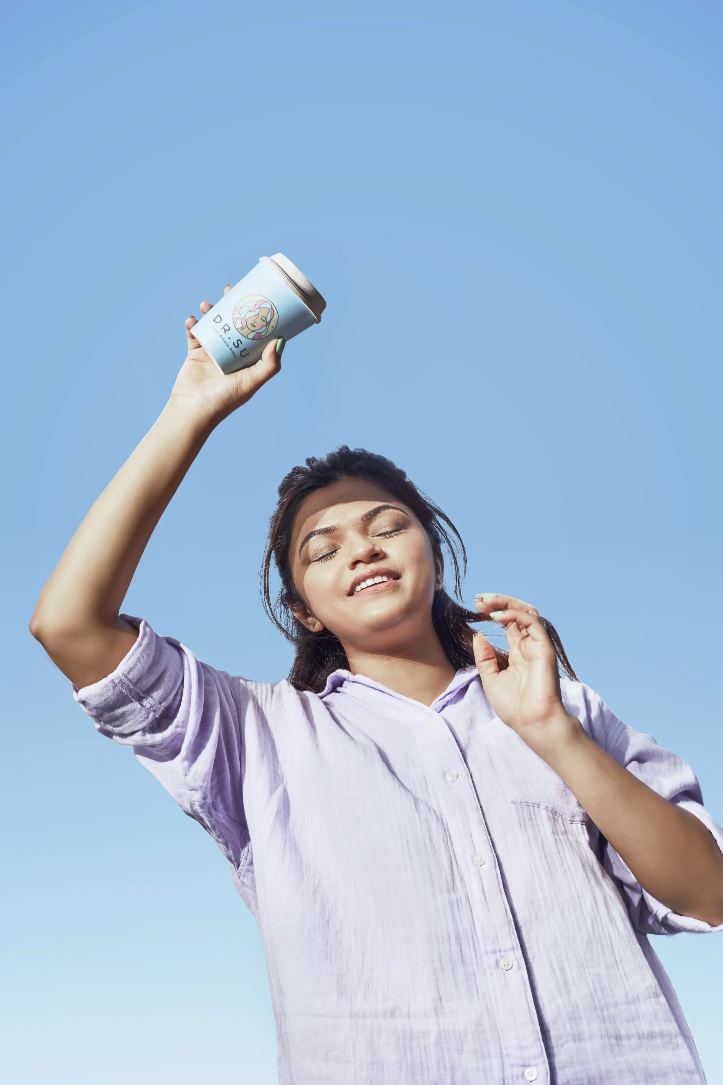 A woman enjoying a sunny day, holding a coffee cup with a colorful cartoon character design against a clear blue sky.
