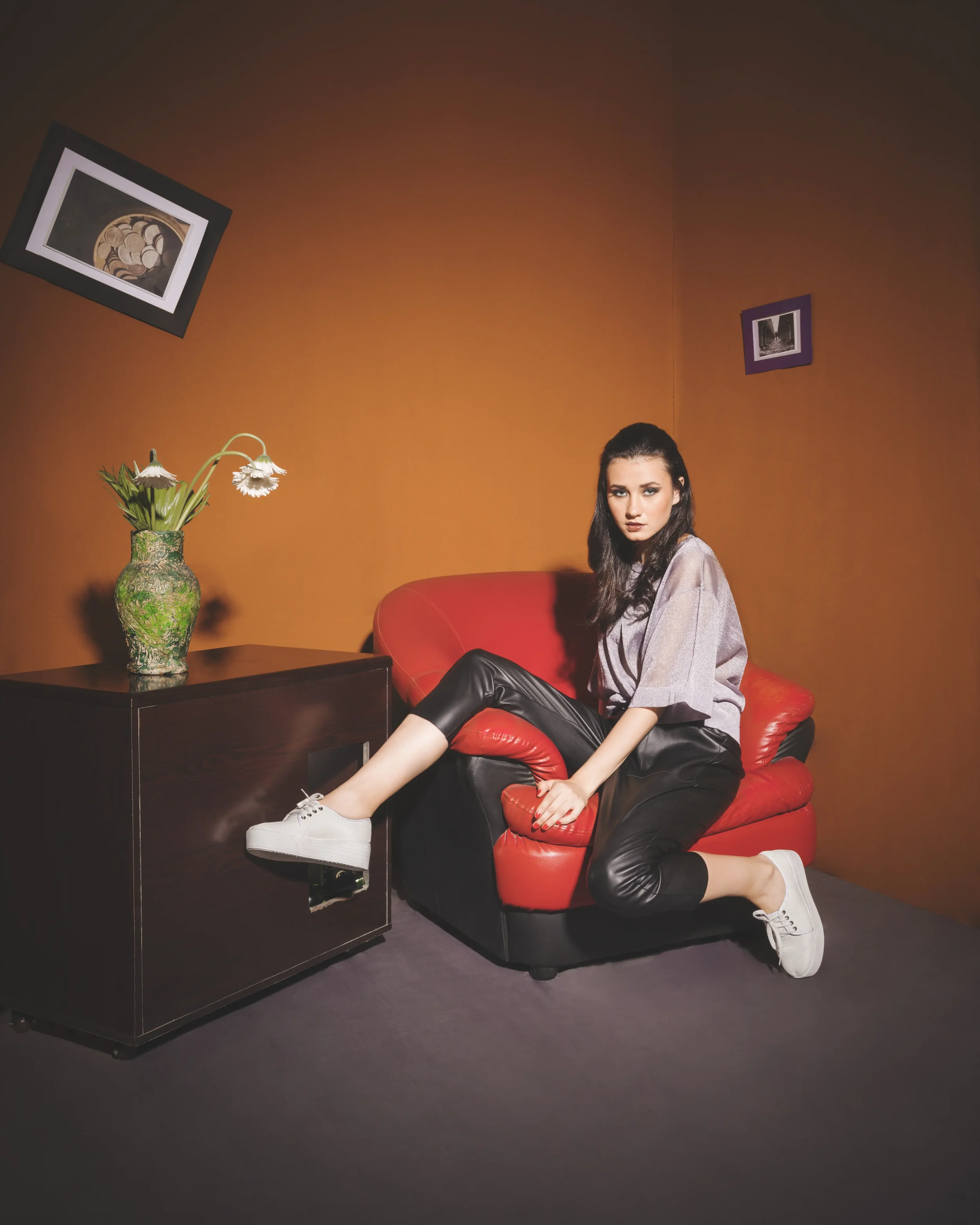 A young woman with dark hair sits on a red leather couch in a room with orange walls. She wears a gray top, black leather pants, and white sneakers. A dark wooden side table with a green patterned vase and white flowers sits beside her. There are two