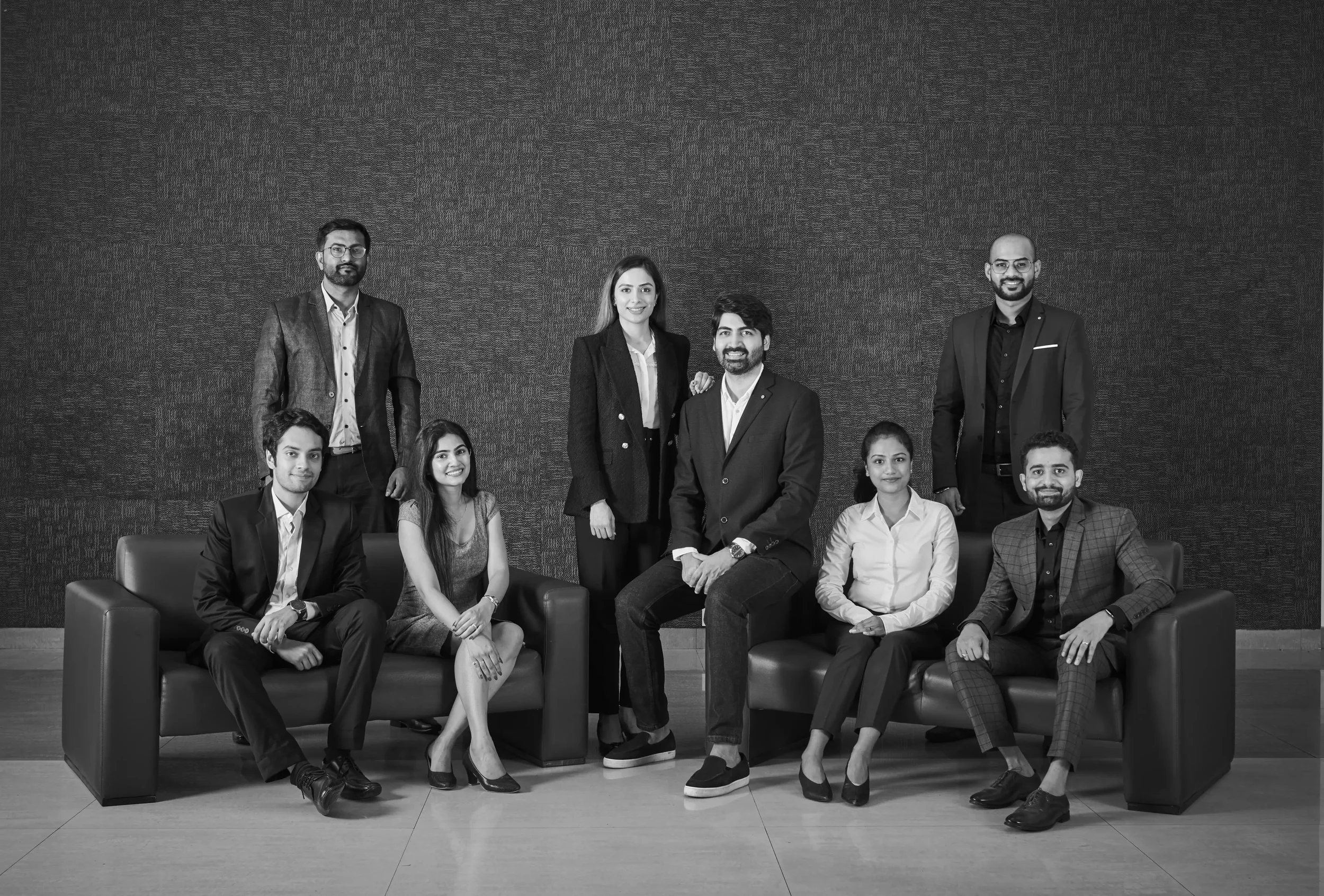 Group of ten professionally dressed people posing for a photo in a modern office or conference room, with some seated and some standing, against a textured wall.