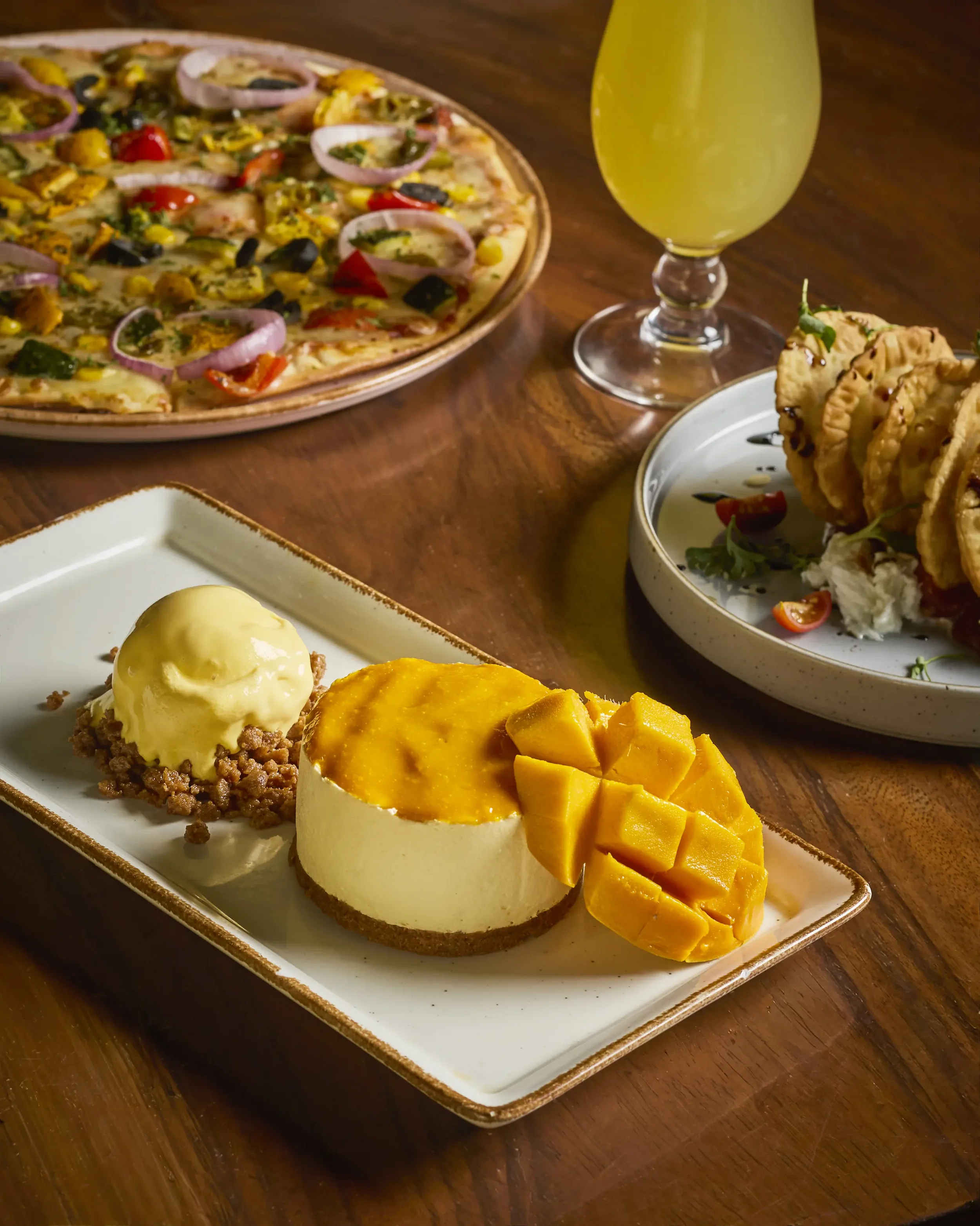 A serving of mango sticky rice, topped with diced fresh mango pieces, served on a rectangular white dish. In the background, a dessert with ice cream and crumbled topping on a white plate, along with a partially visible dessert with a scoop of ice cr