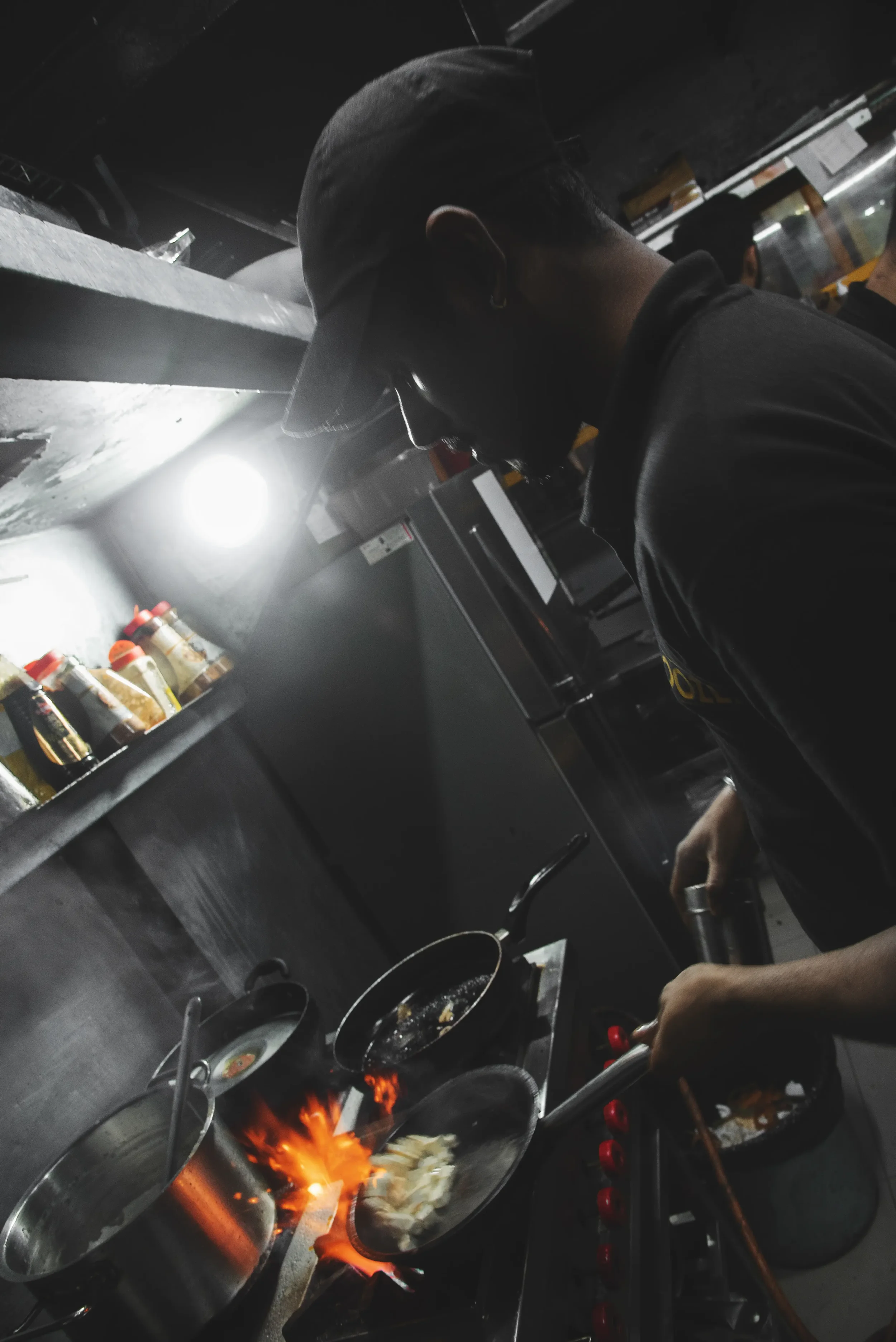 Person cooking on a stove with flames, wearing a baseball cap and dark clothing, in a commercial kitchen.