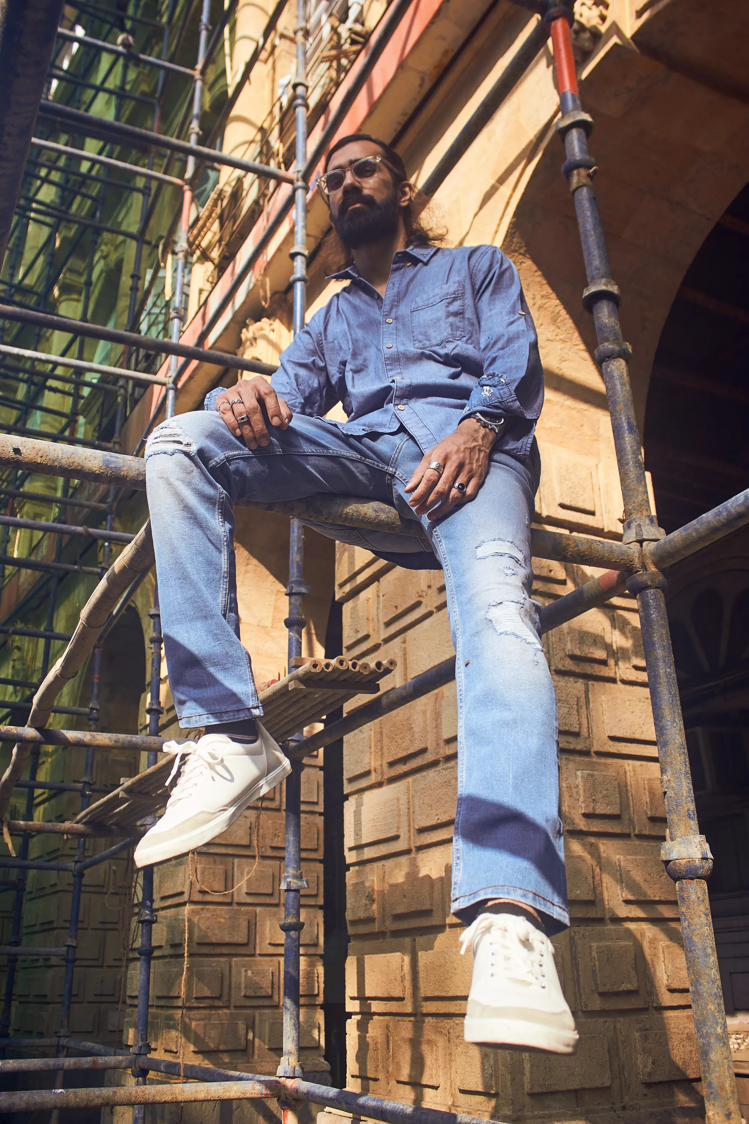 A man with a beard and glasses, wearing a denim shirt, ripped jeans, and white sneakers, is sitting on a metal scaffolding outdoors against a brick building and another building with green windows.