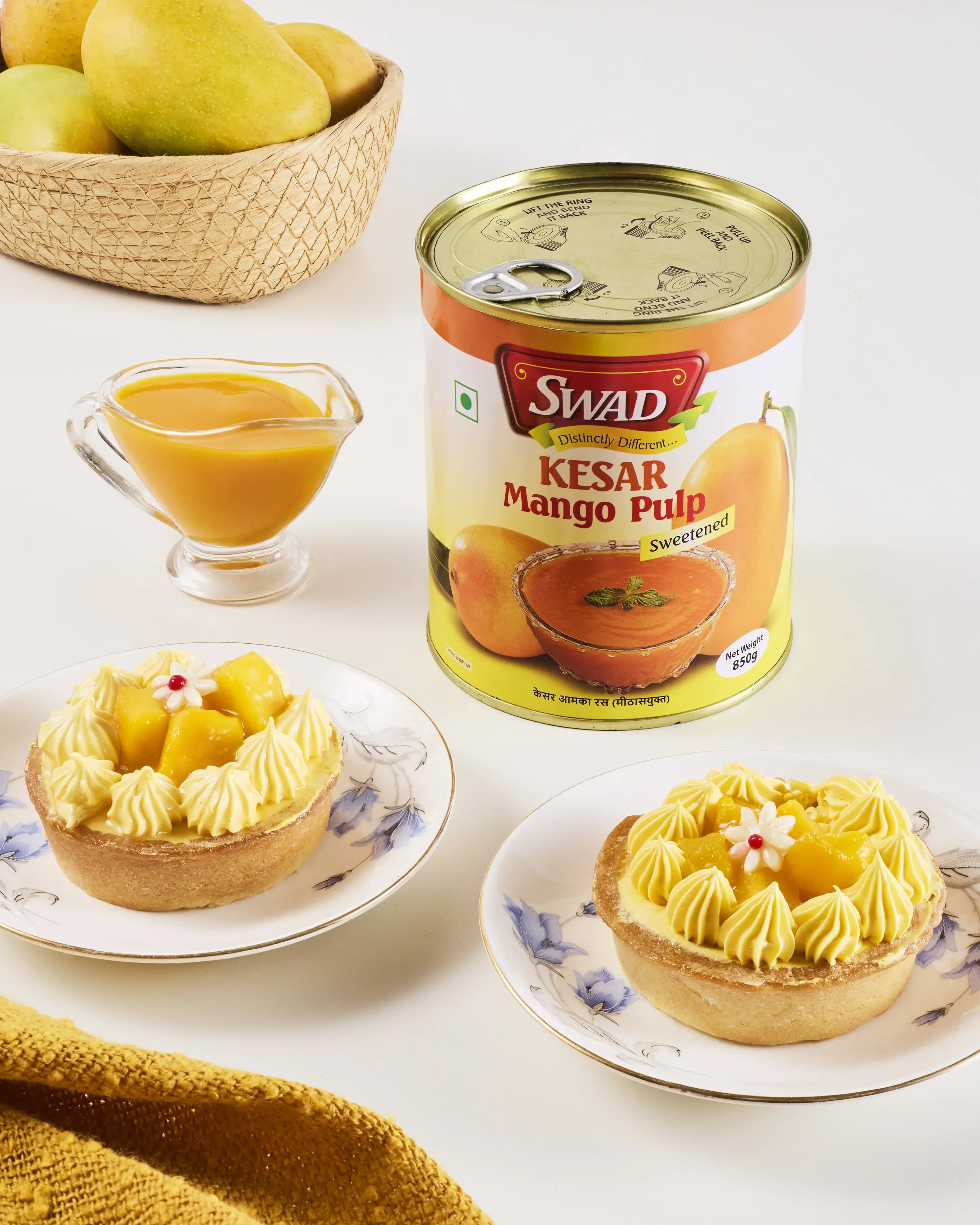 Two fruit tarts topped with mango slices and decorative flowers on floral-patterned plates, a can of Swad Kesar Mango Pulp, a glass pitcher of mango juice, and a basket of apples in the background.