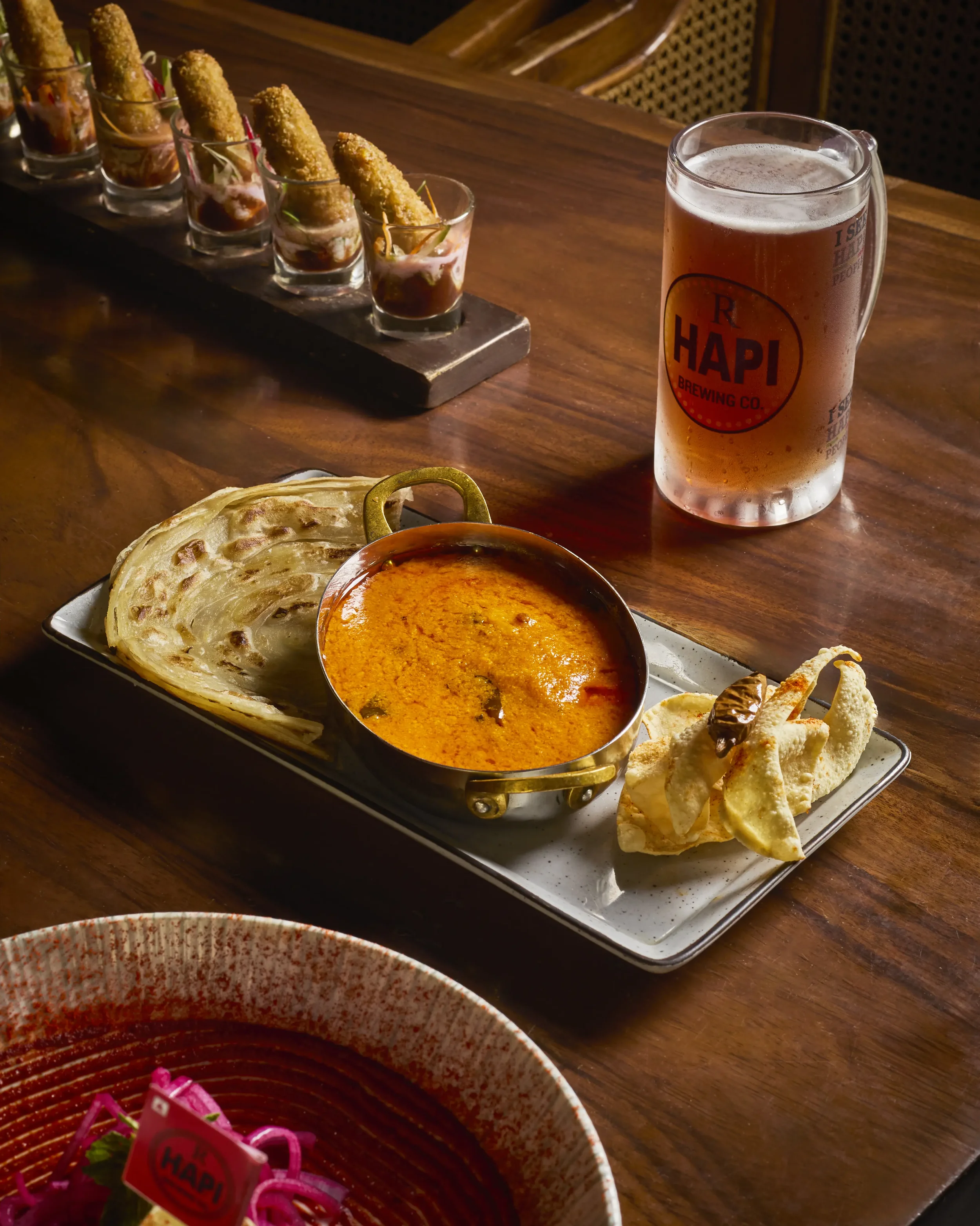 A serving tray containing naan bread, a bowl of Indian curry, and fried papadum chips. In the background, there is a row of shot glasses filled with a layered appetizer or dessert, each garnished with a fried item on top. To the right, a glass of bee