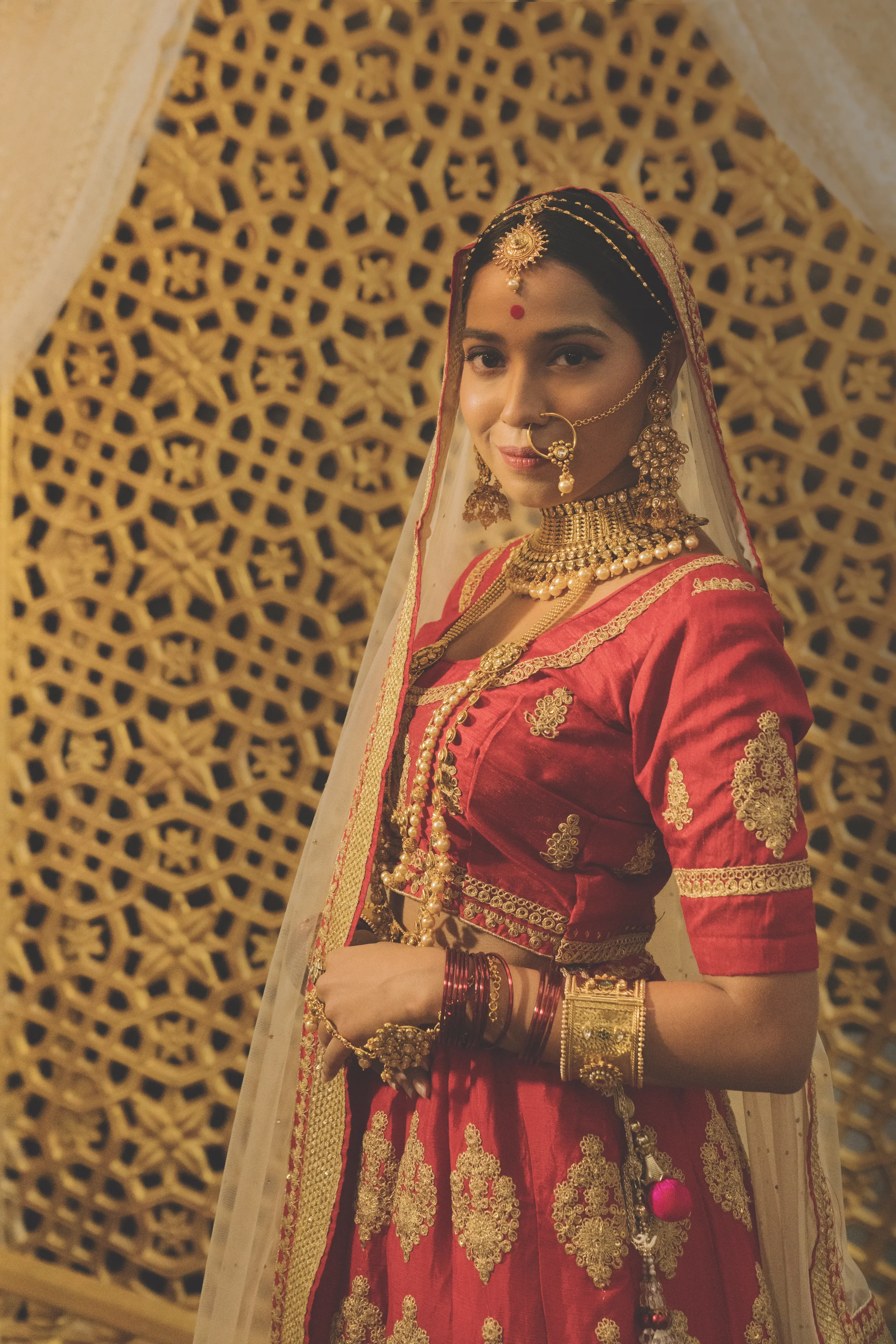 A woman dressed in traditional Indian bridal attire with red and gold embroidery, adorned with gold jewelry including a necklace, earrings, nose ring, and bangles, standing in front of a decorative golden lattice background.