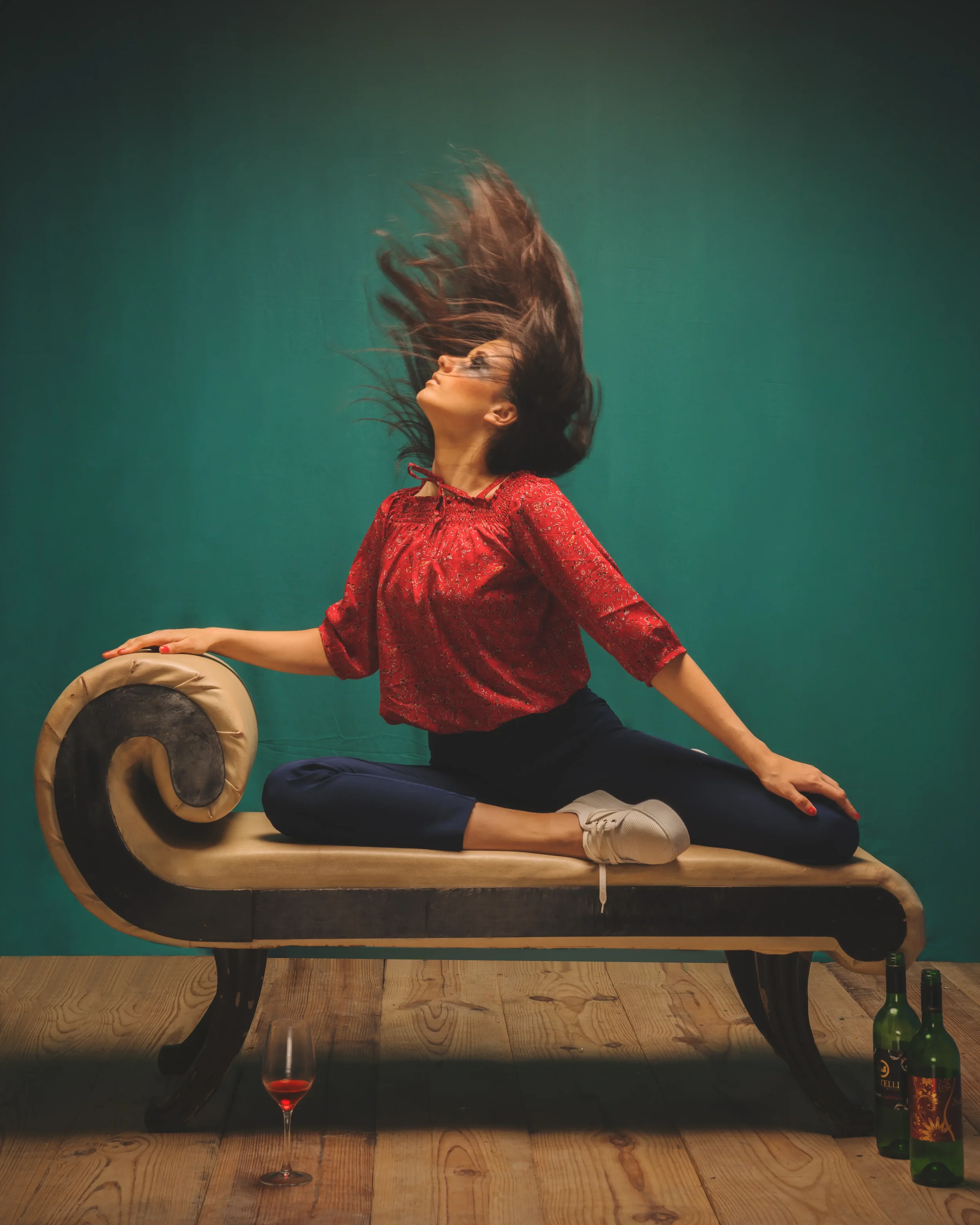 A woman in a red blouse and dark pants sitting on a vintage chaise lounge with her hair flying, against a green background. There are two bottles and a wine glass on the floor.
