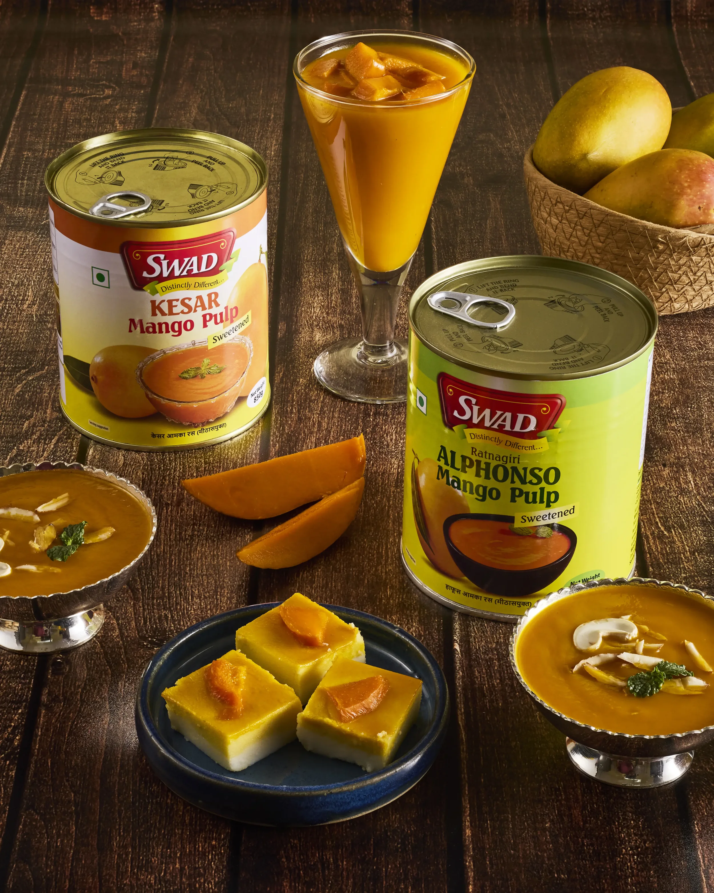 A table with canned mango pulp, a glass of mango pulp with pieces of mango, slices of mango, and Indian dessert Kalakand garnished with pistachios and mint leaves.
