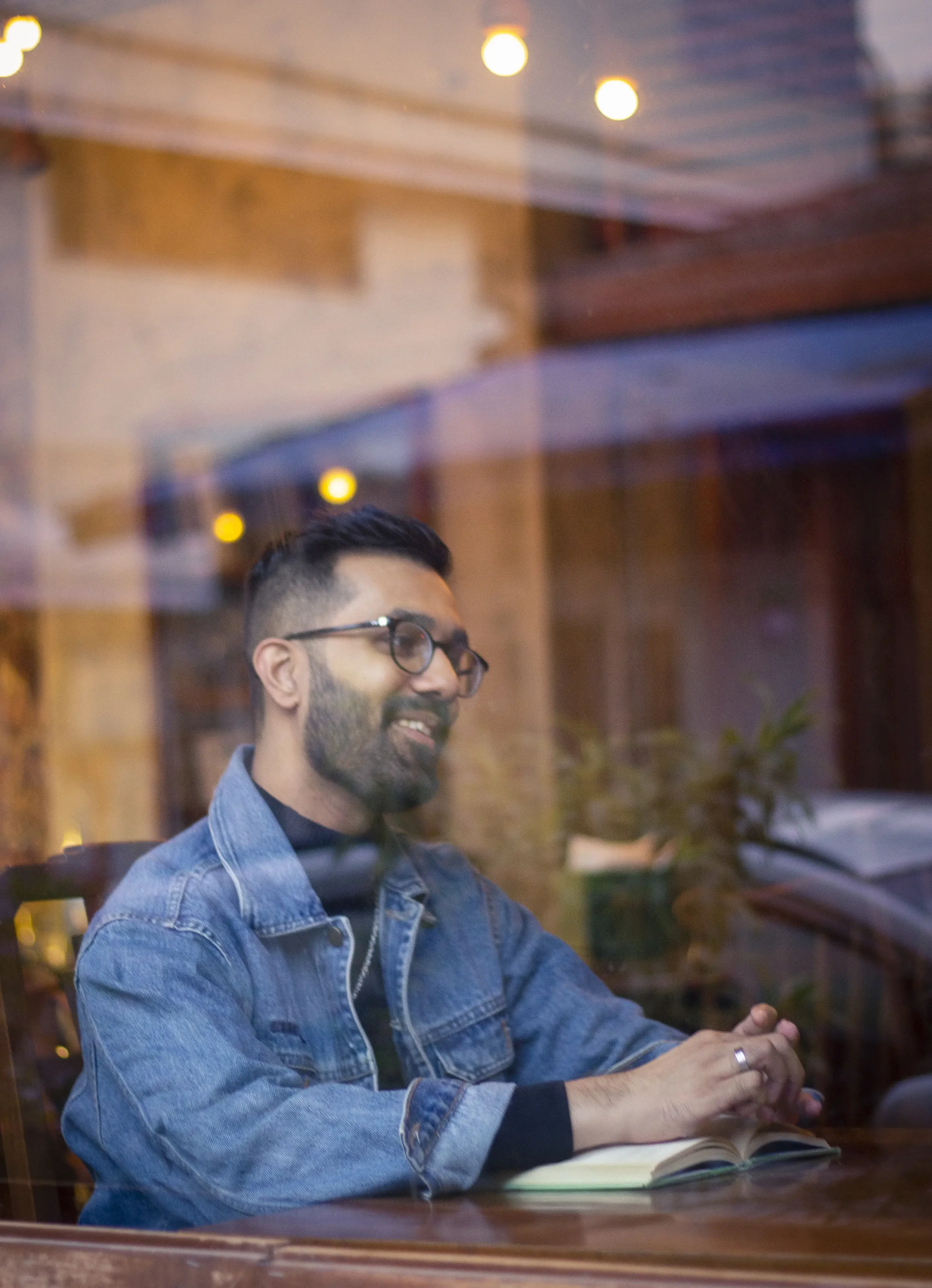 A man with glasses, a beard, and short dark hair sitting at a table, smiling, and looking through a window.