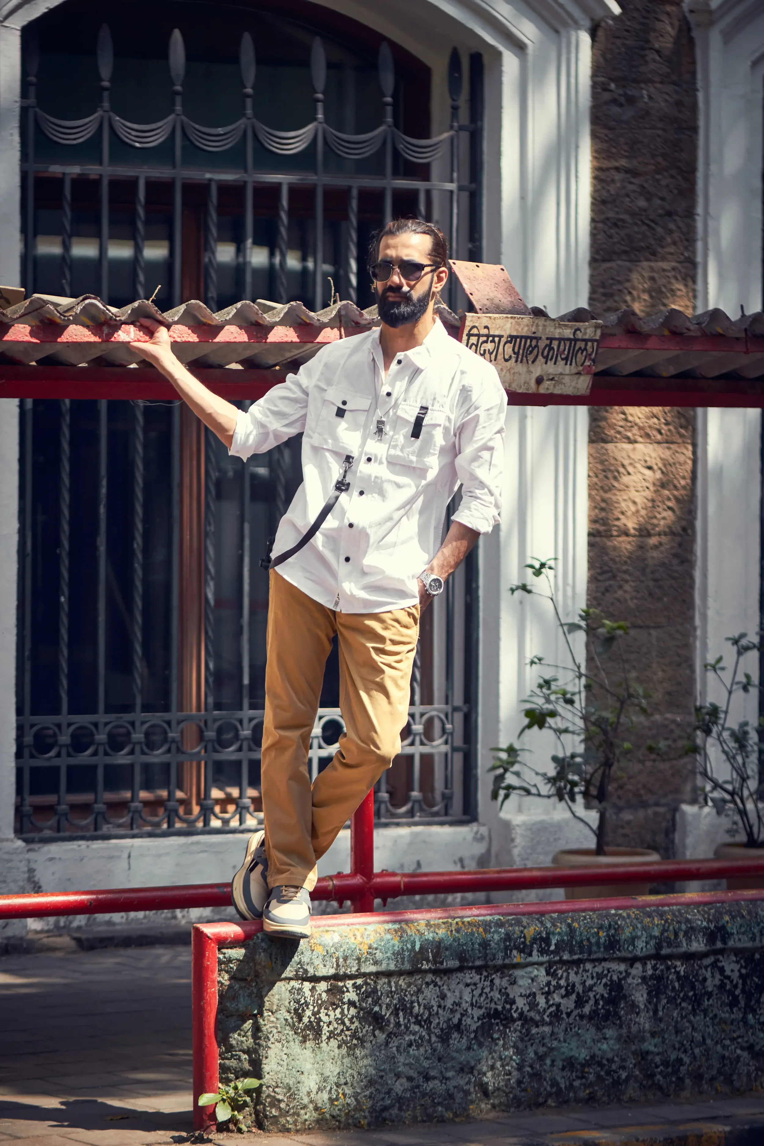 A man wearing sunglasses, a white button-up shirt, and tan pants, standing on a concrete barrier with a red railing in front of a building with barred windows, and holding the railing with one hand.