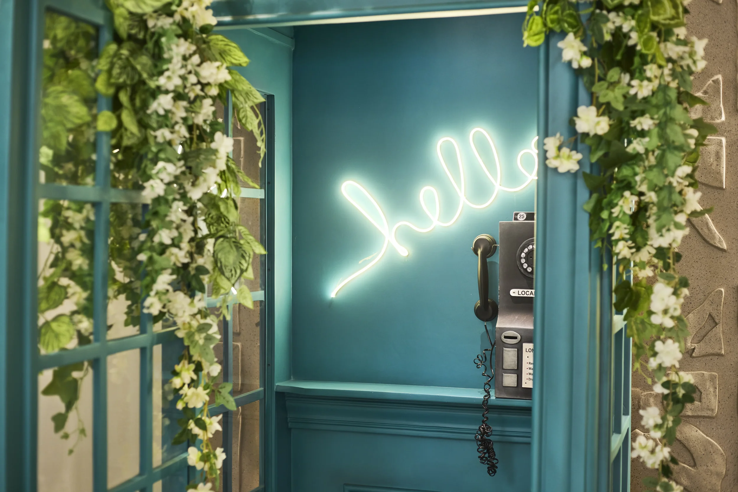 An empty teal-colored phone booth decorated with green and white artificial flowers, with a neon sign spelling 'Hello' on the back wall, and a black payphone attached inside.