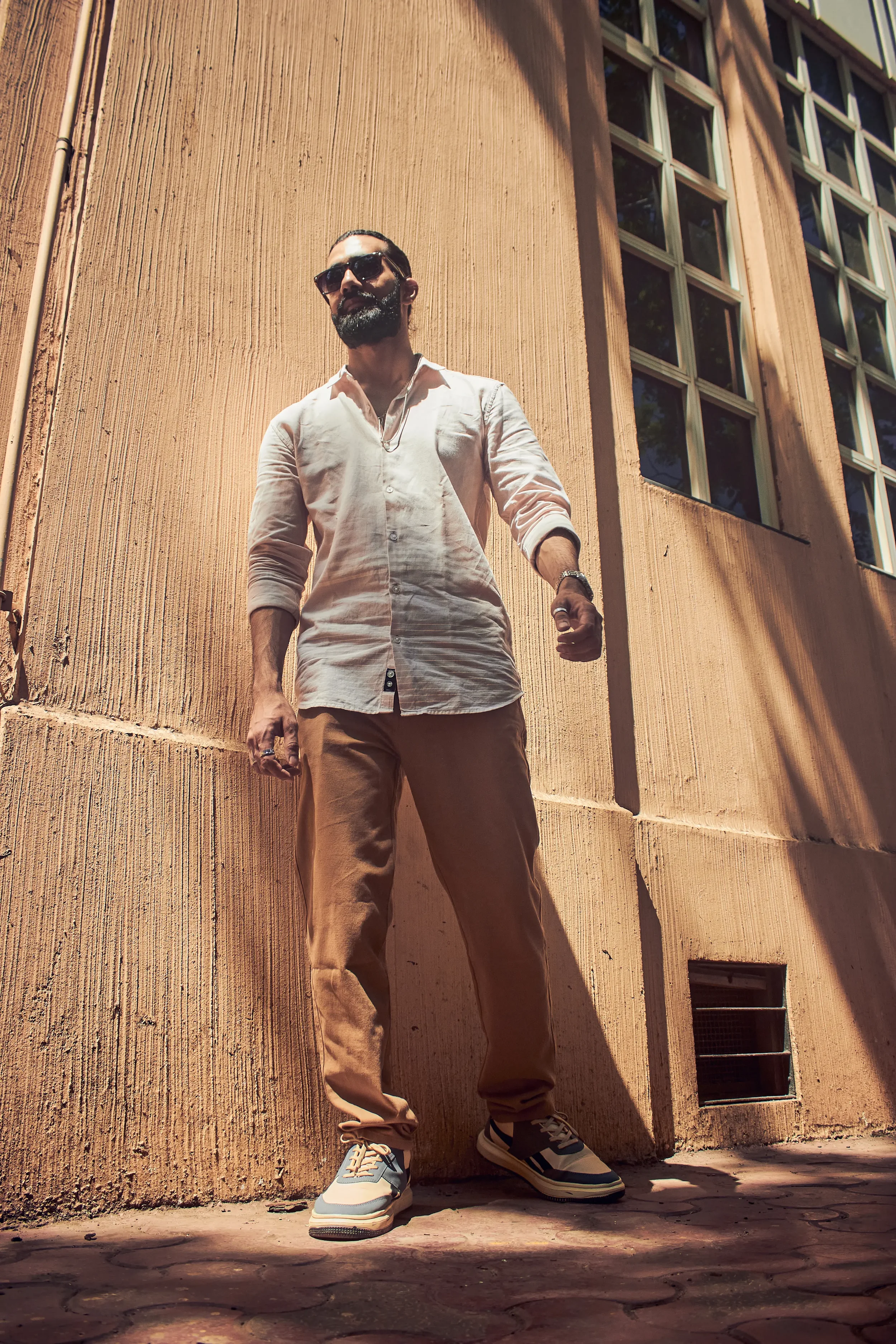 A man with sunglasses, a beard, and dressed in a white shirt and beige pants stands outdoors against a textured beige wall with large windows, looking confident.
