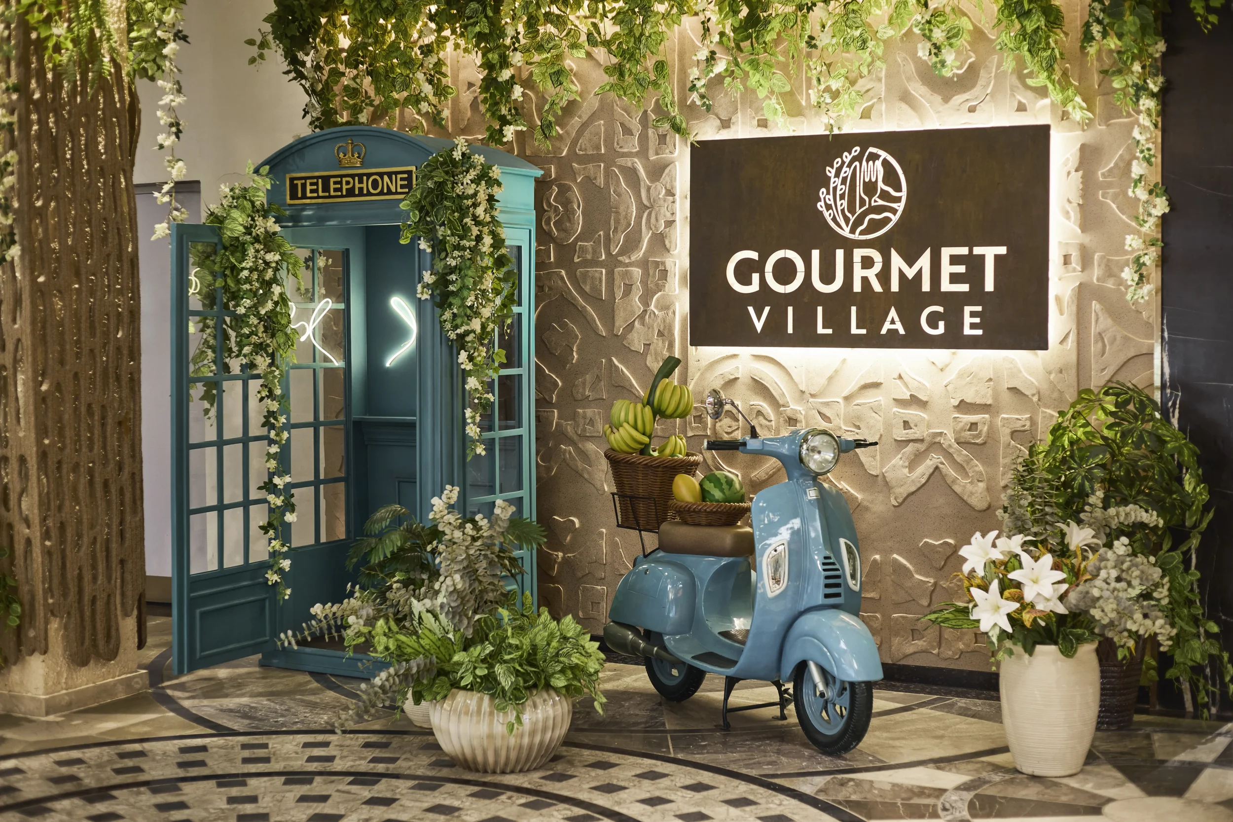 Decorative indoor scene with a blue vintage payphone booth decorated with white flowers and green foliage, a small vintage blue scooter with a basket of green and striped melons, and a large sign reading 'GOURMET VILLAGE' on a textured beige wall wit