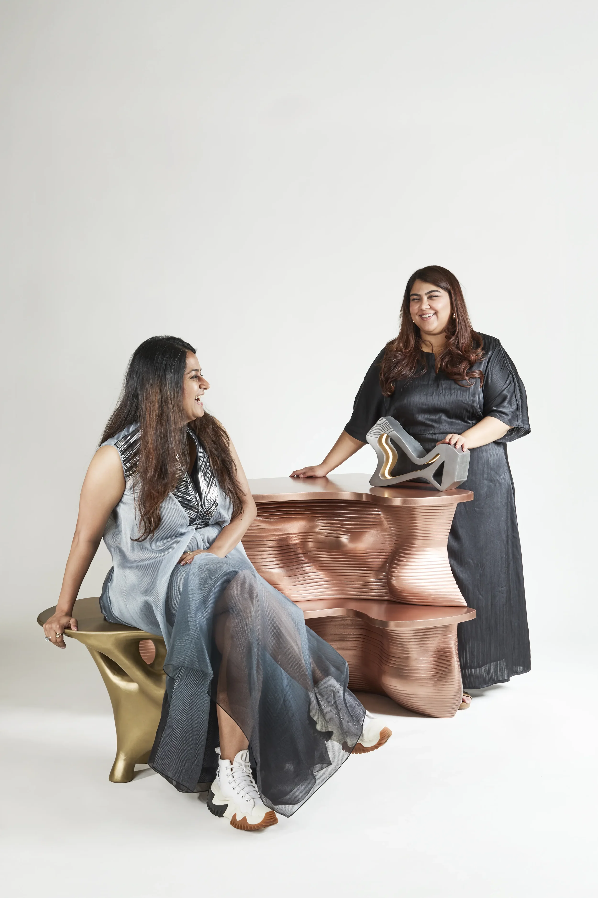Two women with long brown hair, one seated and the other standing, smiling and having a conversation in a minimalist studio with modern furniture and art decor.