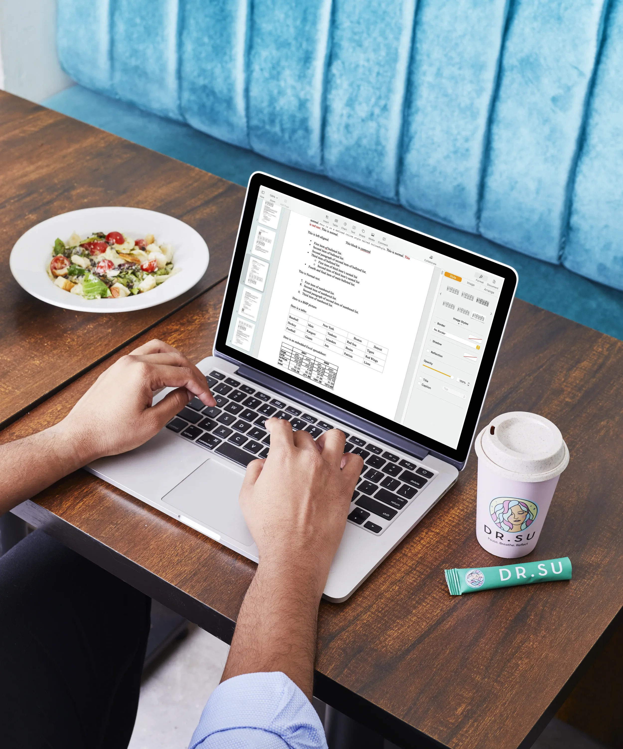 Person working on a laptop at a wooden table, with a salad, coffee, and a tube of Dr. Su intellectual property cream nearby.