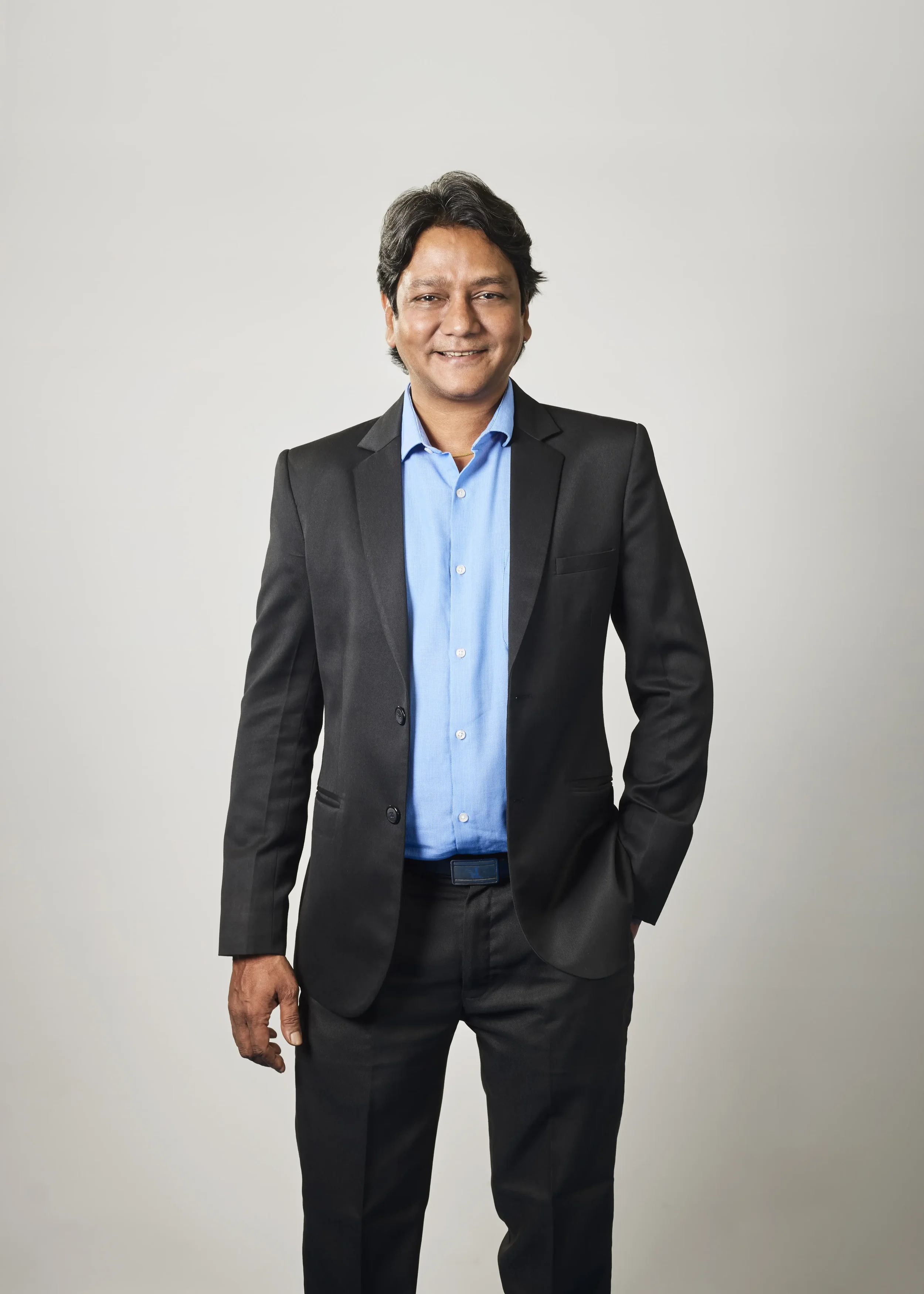 Smiling man in a black suit and light blue dress shirt standing against a plain light grey background.