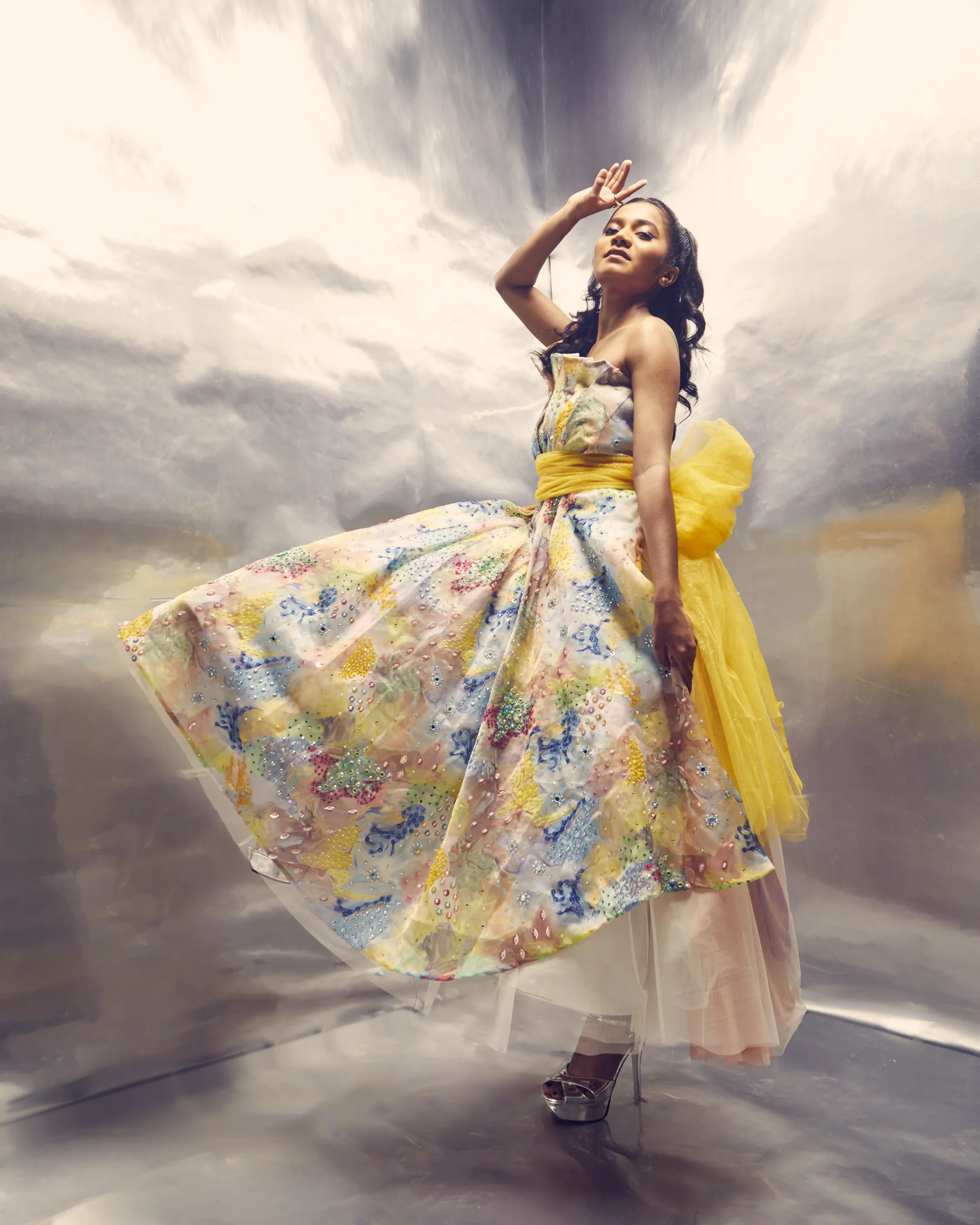 A woman in colorful, floral, and pastel costume with yellow accents, standing in front of a reflective, metallic backdrop, striking a confident pose with one hand near her head and the other palm facing outward.