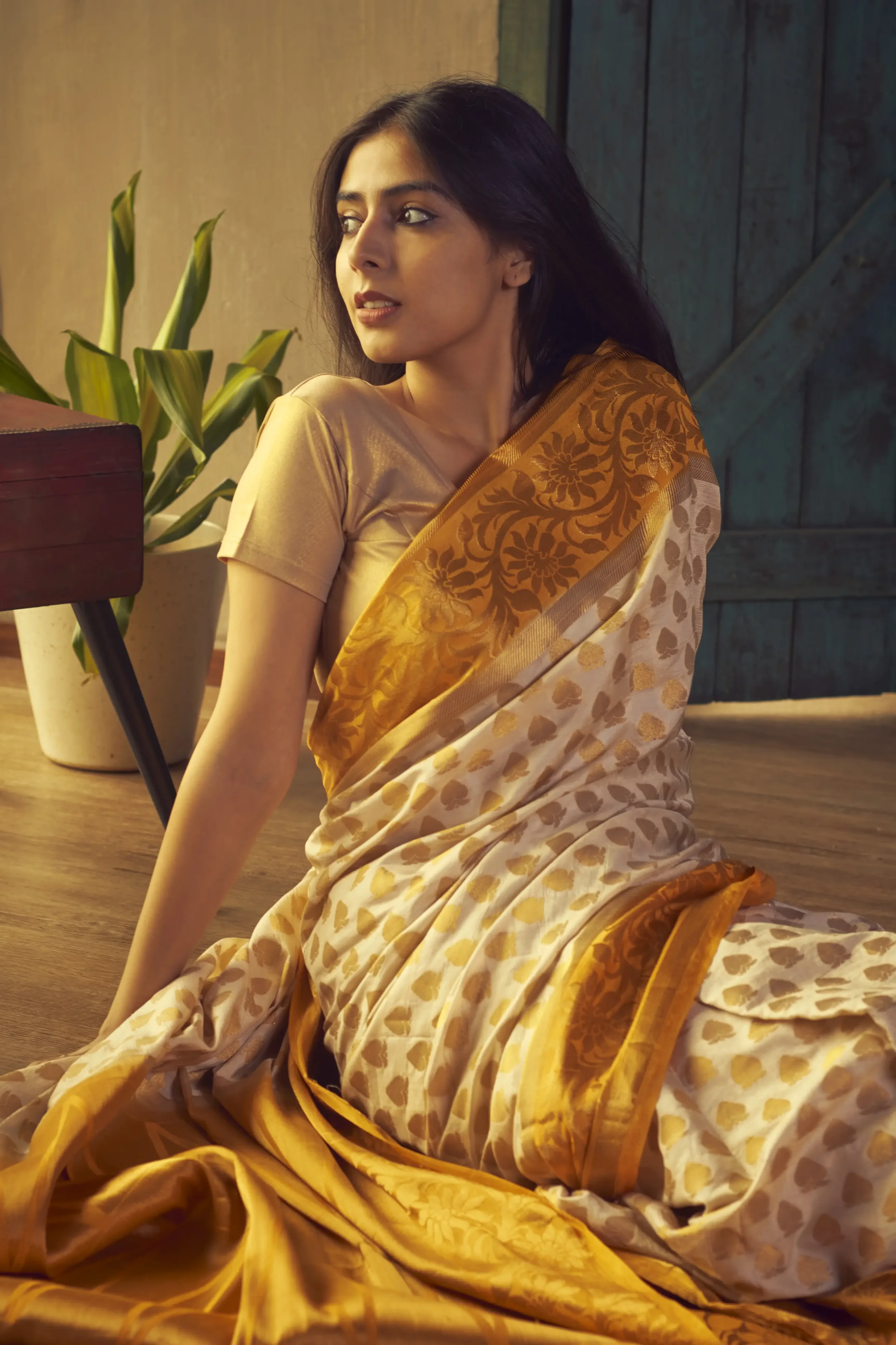 A woman in a gold and cream-colored sari sitting on a wooden floor next to a potted plant, looking to her left with a pensive expression.