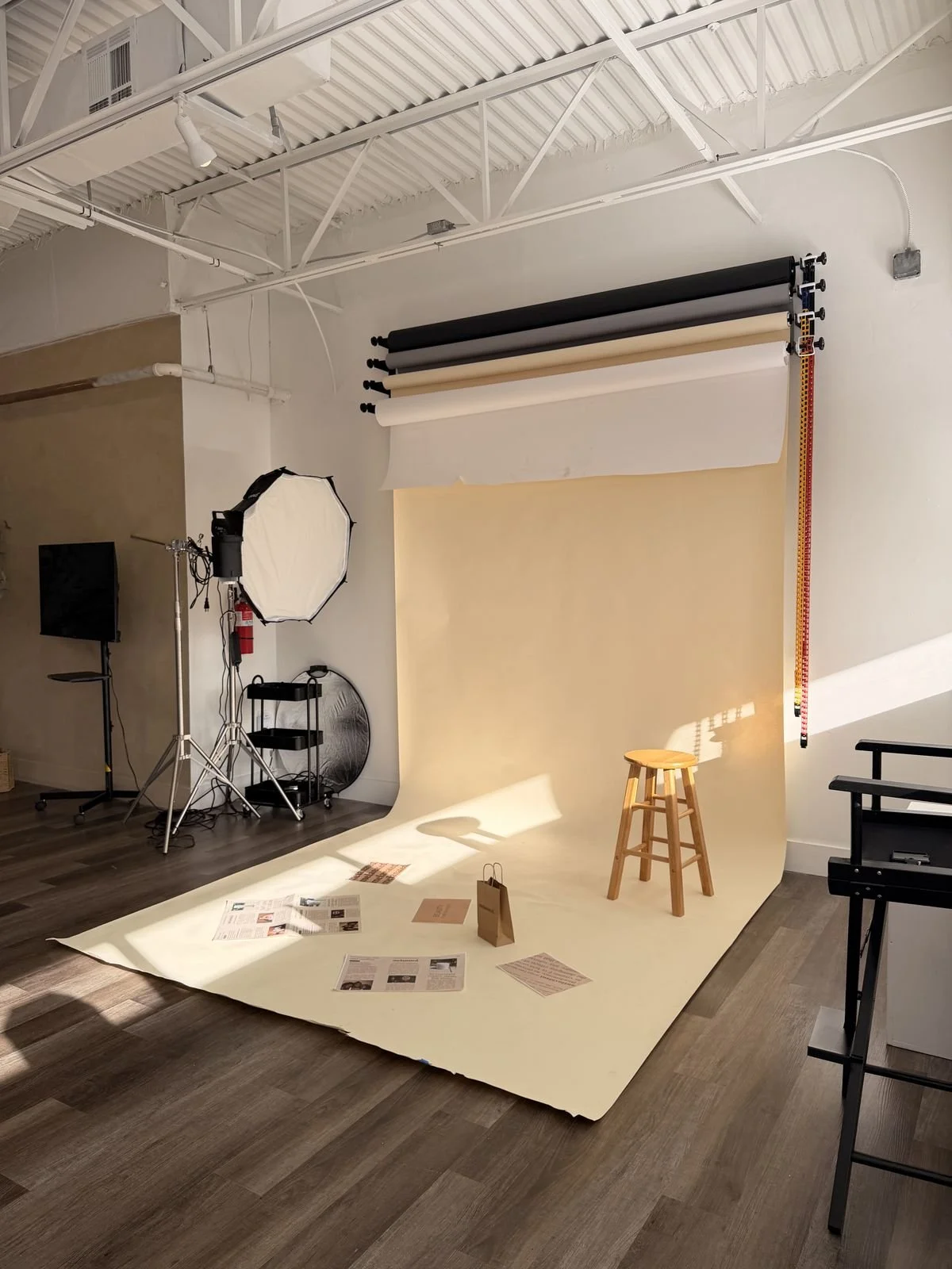 Photography studio with a beige backdrop, stool, and lighting equipment including a large softbox, background rollers, and papers and magazines on the floor.