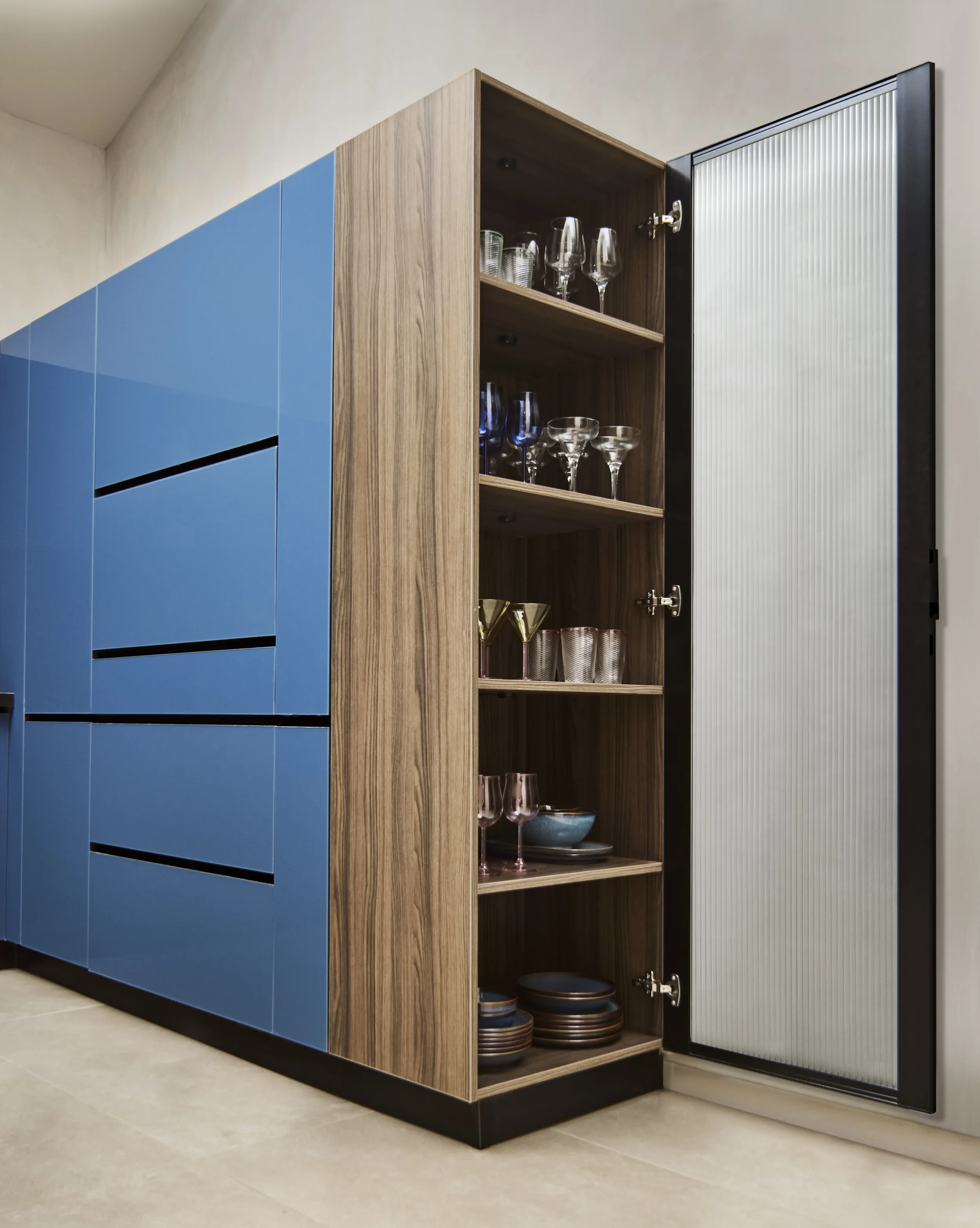 A modern kitchen cabinet with blue doors on the left and a wood-paneled section on the right, with open shelves containing glasses, bowls, and dishes. The cabinet has a black and white slatted door on the right side.