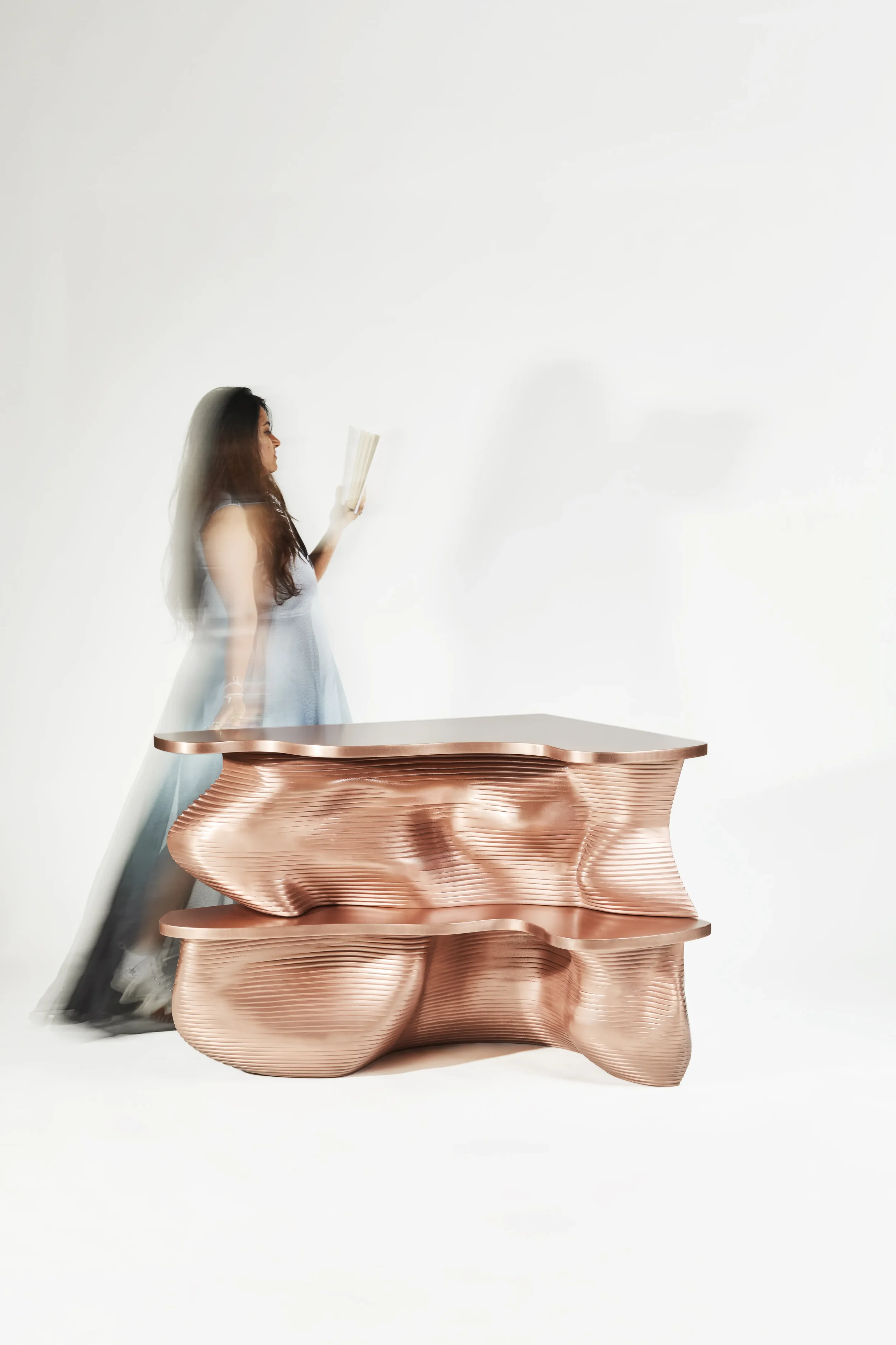 A woman in a light blue dress stands next to a modern, abstract copper-colored table or sculpture, holding a book and facing in profile against a plain white background.