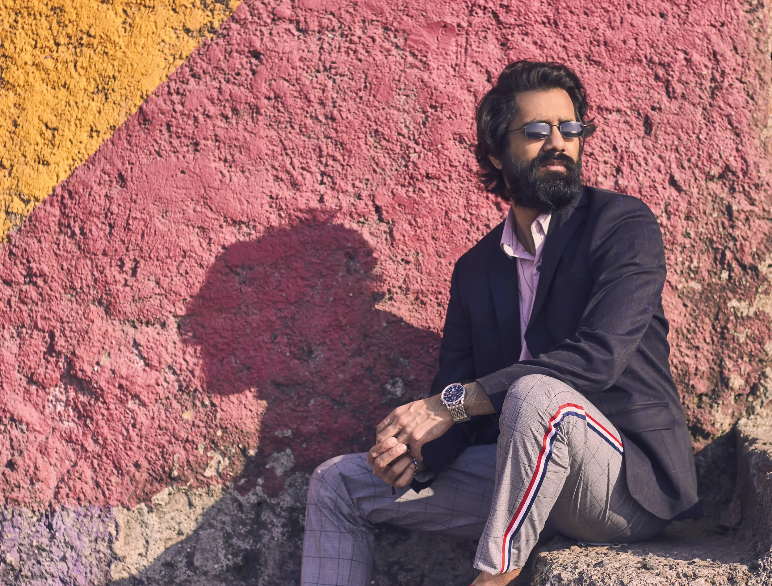 A man sitting on rocks in front of a pink and yellow painted wall, wearing sunglasses, a black blazer, a light shirt, checkered pants with red, white, and blue stripes, and a watch.