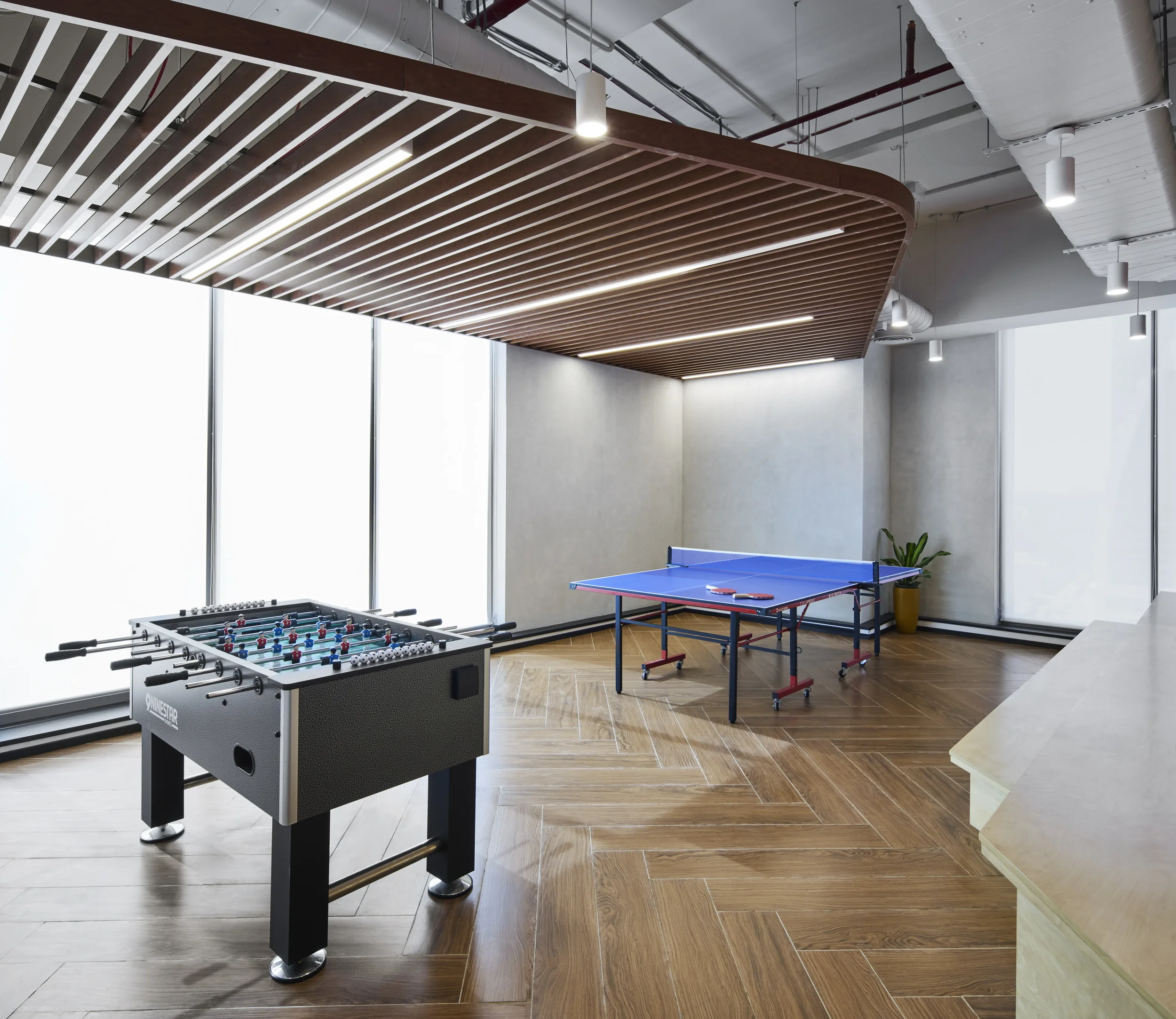 An indoor recreational space featuring a foosball table in the foreground, a ping pong table to the right, and a modern ceiling with metal ducts and ceiling lights. Large windows provide natural light.