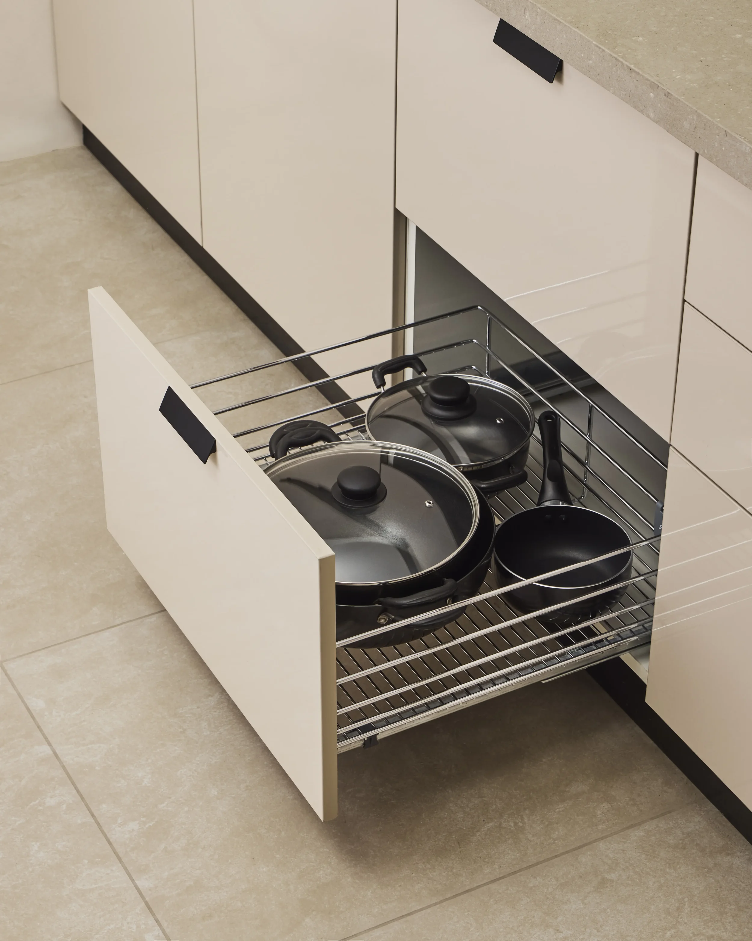 Open kitchen drawer with pots and pans inside, located beneath a beige countertop and against beige cabinets on a beige tile floor.