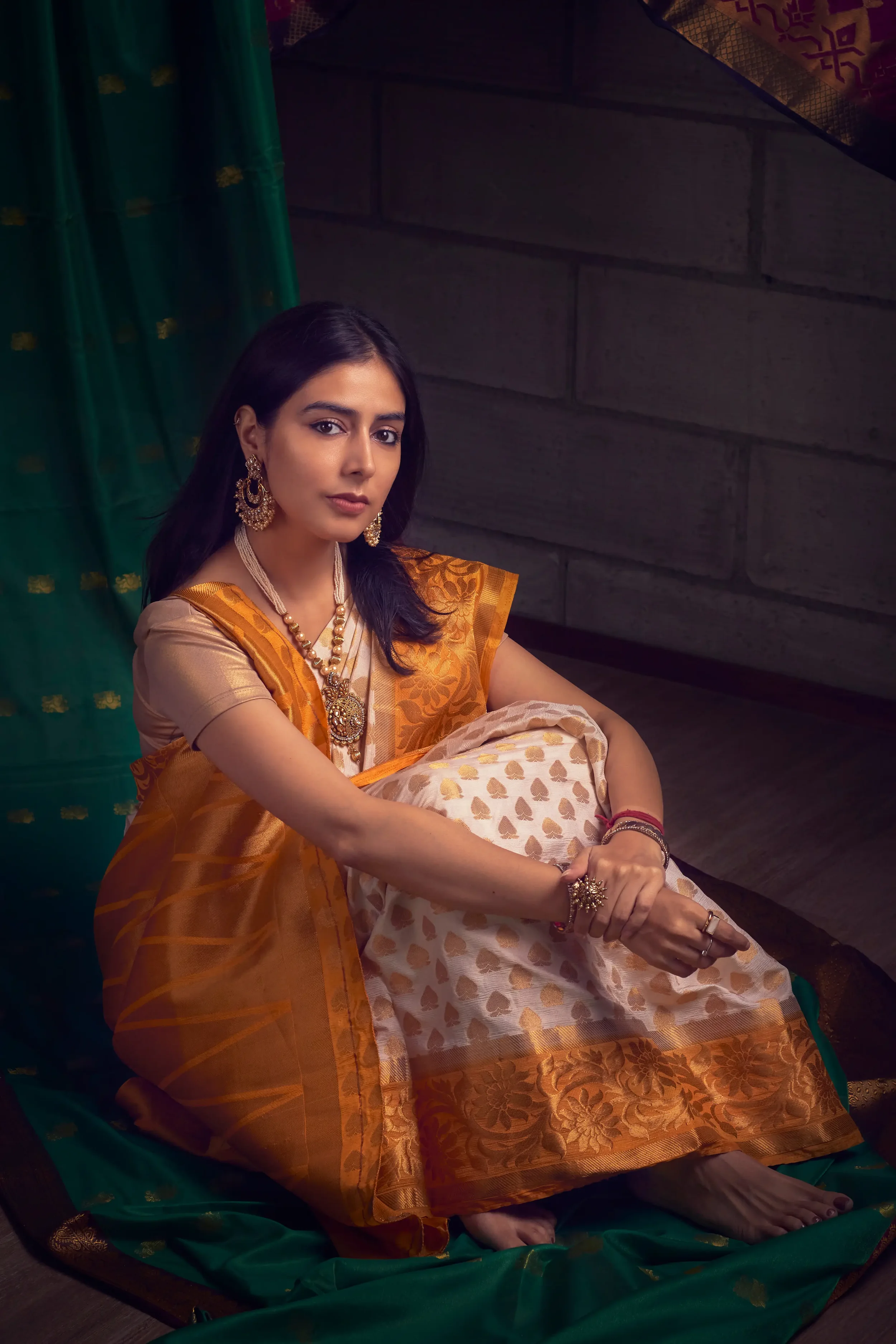 A woman dressed in traditional Indian attire, including a golden and orange saree with intricate designs, sitting on a green cloth. She has long dark hair and is wearing gold jewelry, including earrings, necklaces, and bangles, with a neutral facial 