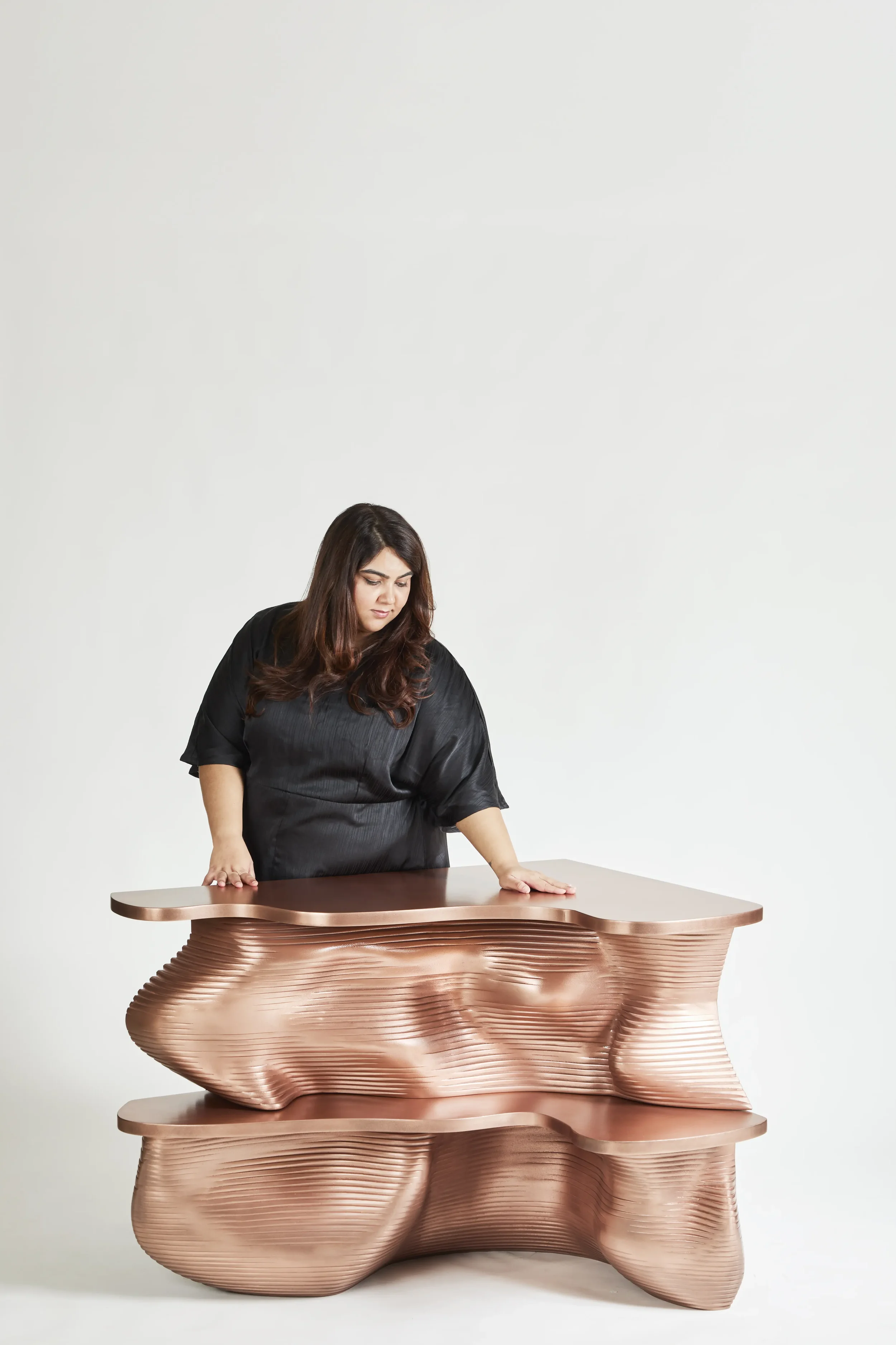 A woman in black clothing leaning on a modern, abstract copper-colored sculptural table.