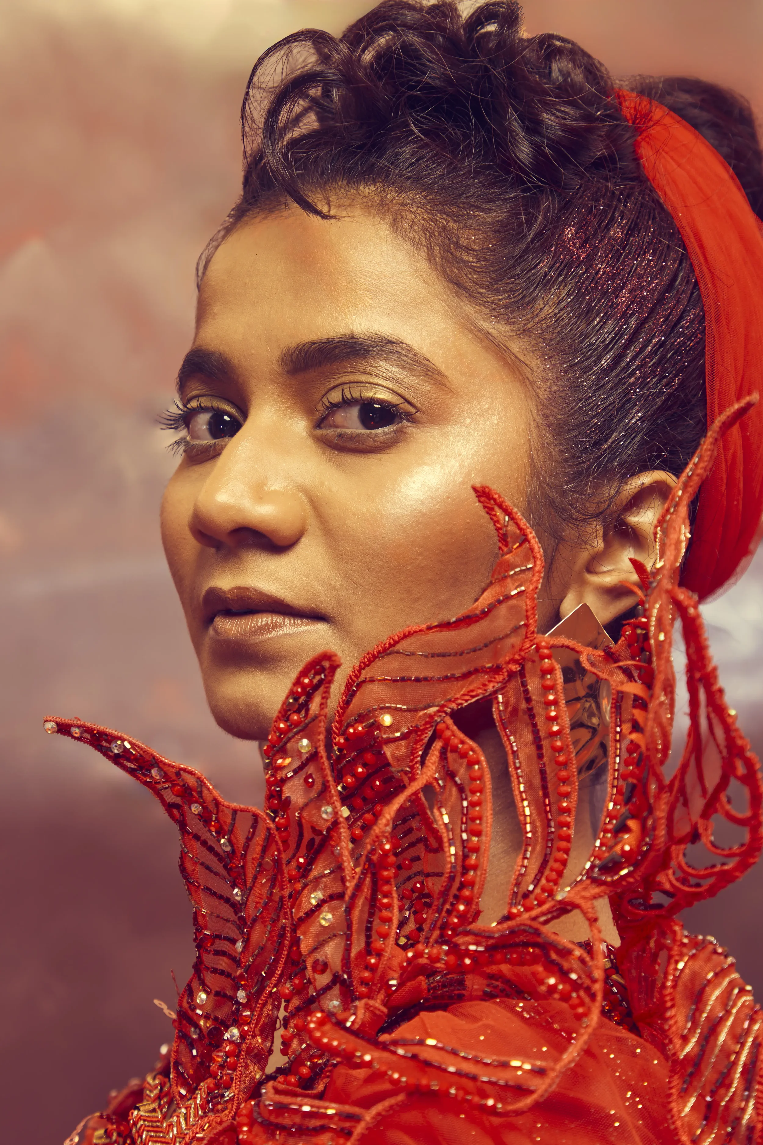 Close-up of a woman with dark hair, wearing red headband and an ornate red and gold garment with intricate embroidery and beadwork.