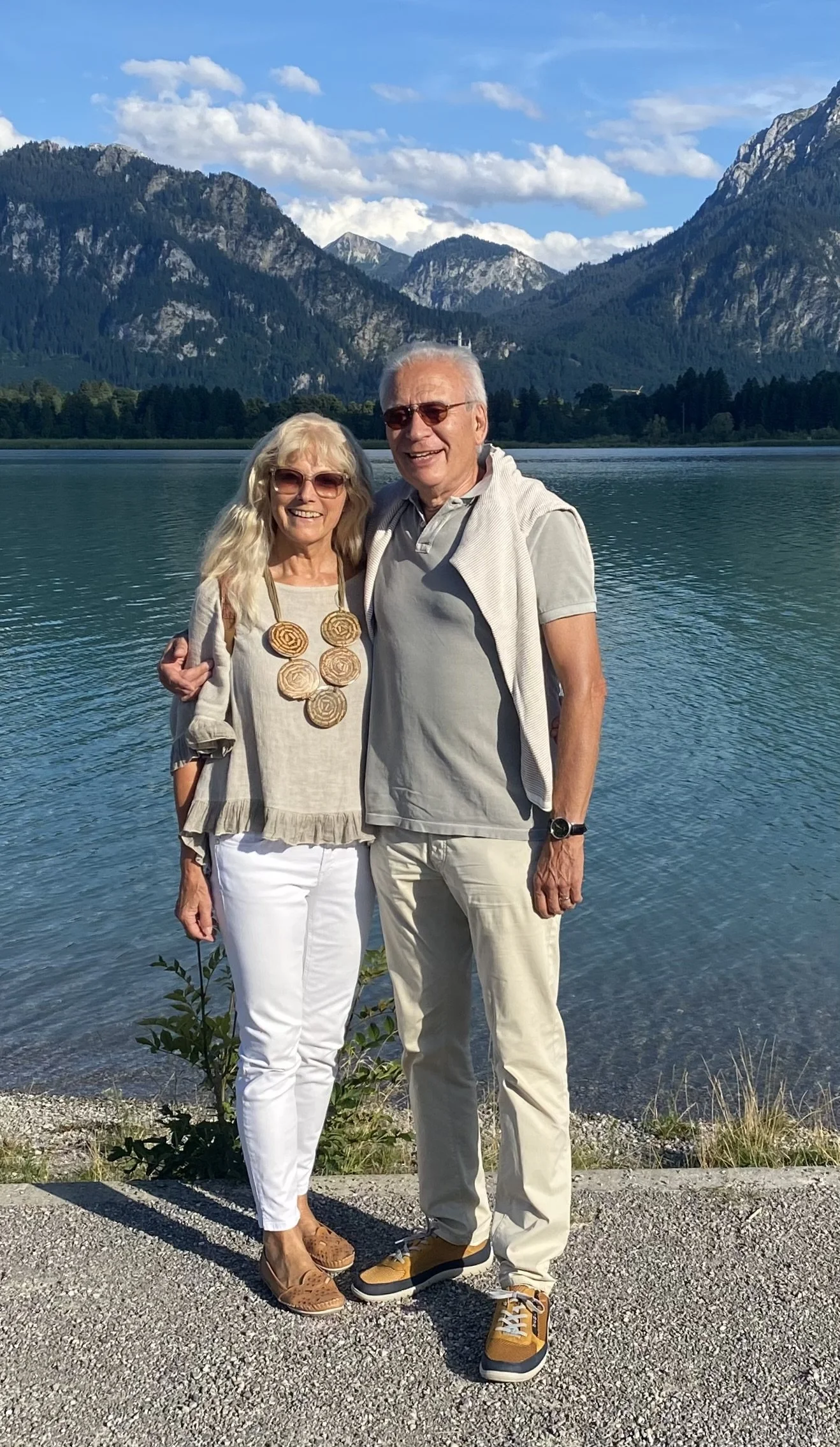 Ein freundliches Ehepaar steht an einem See mit Bergen im Hintergrund, lächelnd und in Freizeitkleidung.