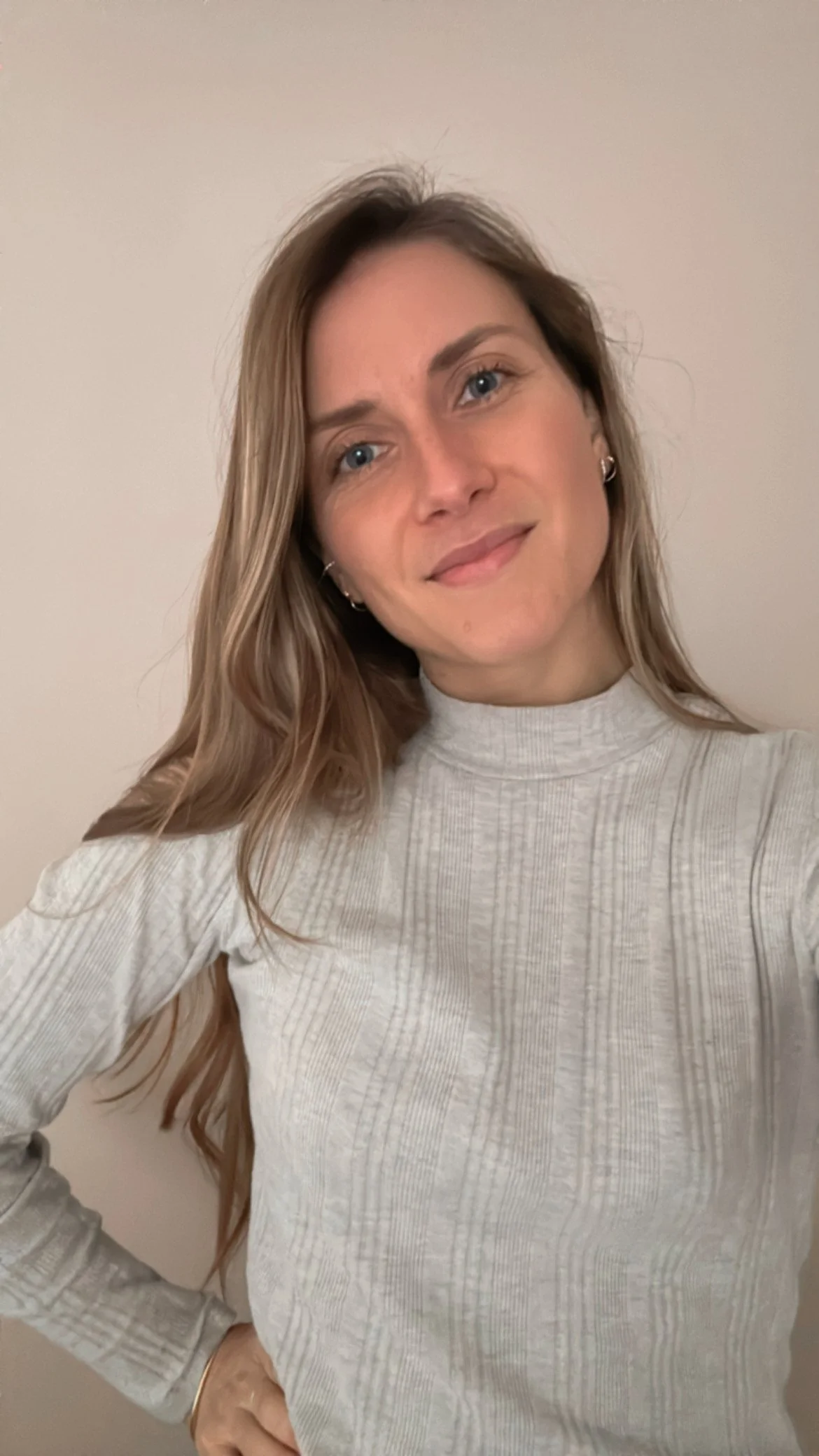 Jeune femme avec des cheveux longs, porter un pull gris à col roulé, souriant, les mains sur les hanches, debout contre un mur blanc.