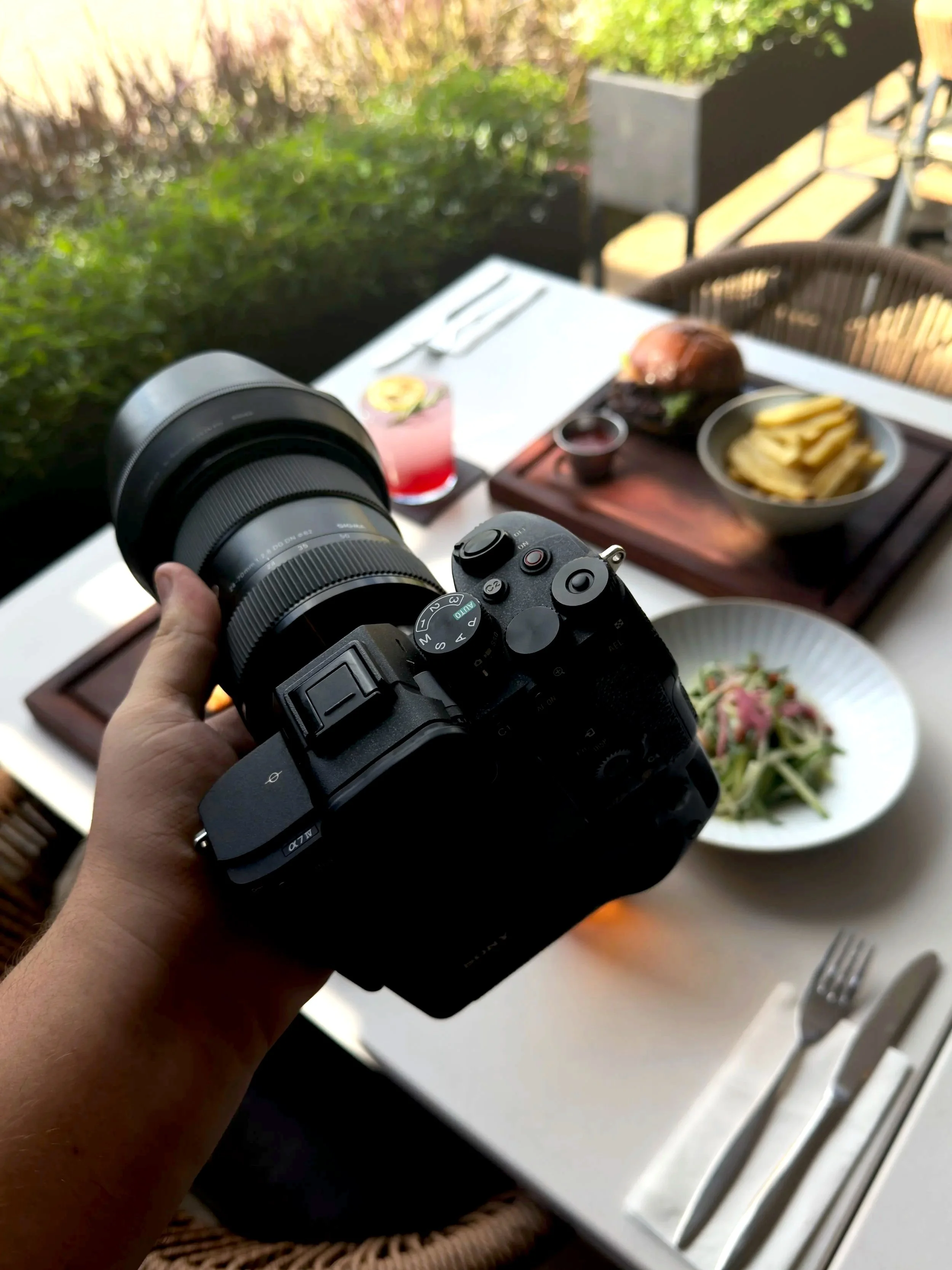 Camera held above a table with a burger, fries, salad, and a drink, set outdoors on a patio.