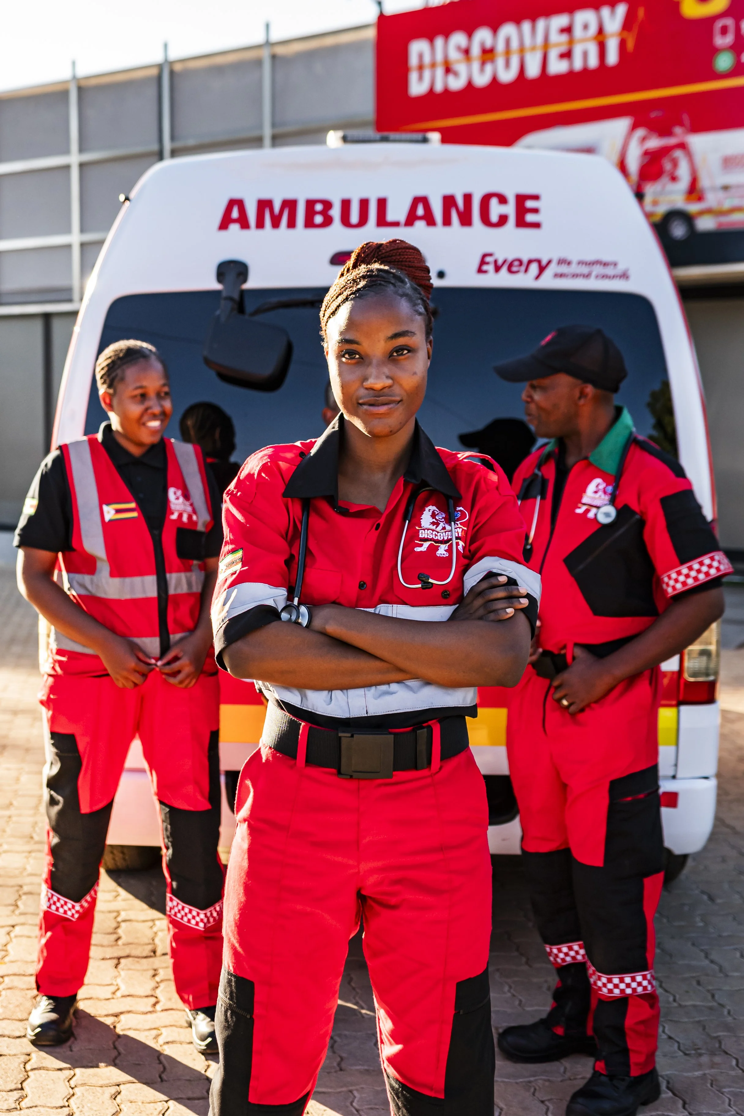 Three emergency medical responders in red and black uniforms standing in front of an ambulance, with a Discovery billboard in the background.