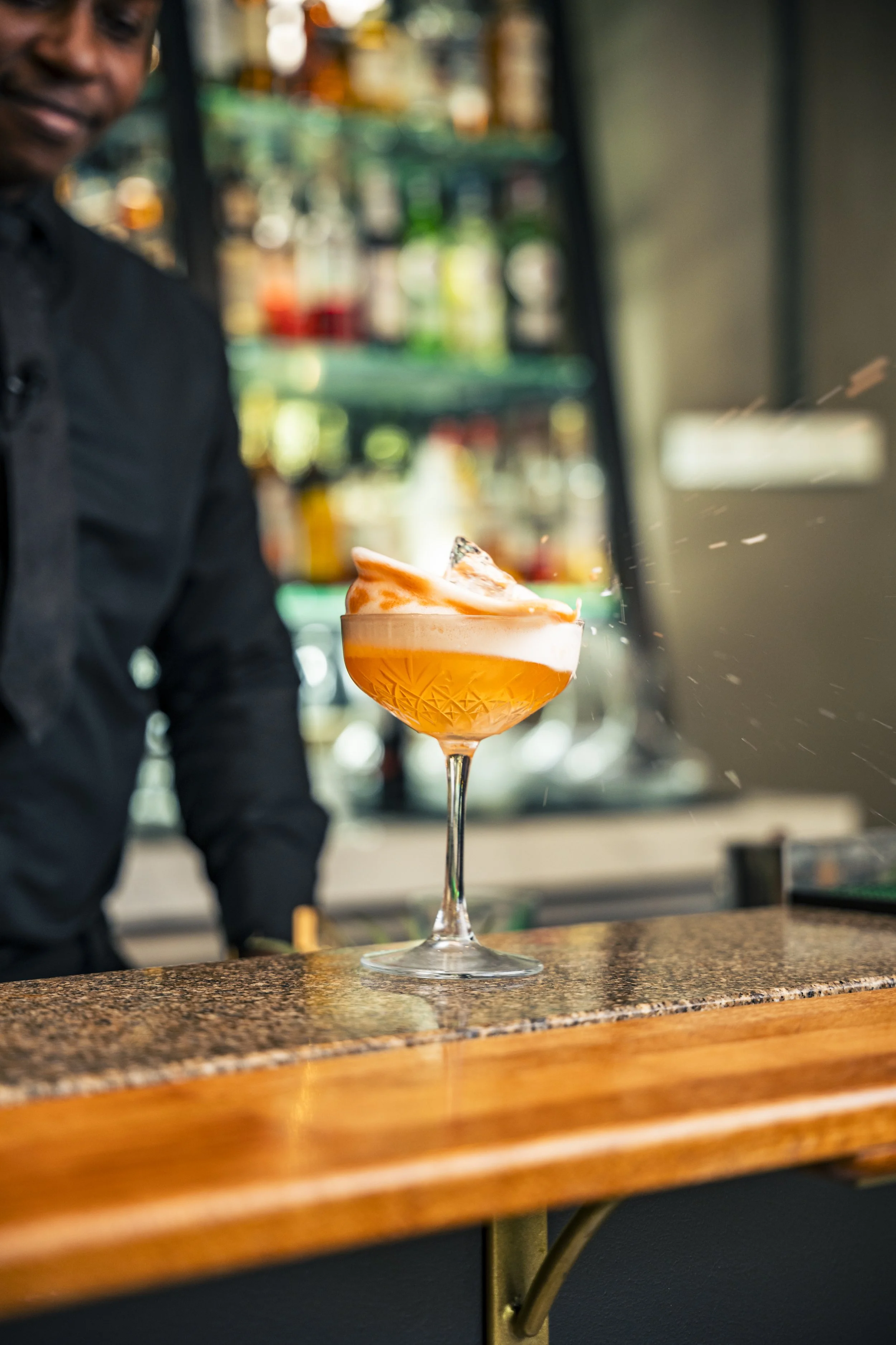 A bartender mixing a cocktail in a glass with orange hue, foam, and garnished with a lemon peel, in a bar setting.