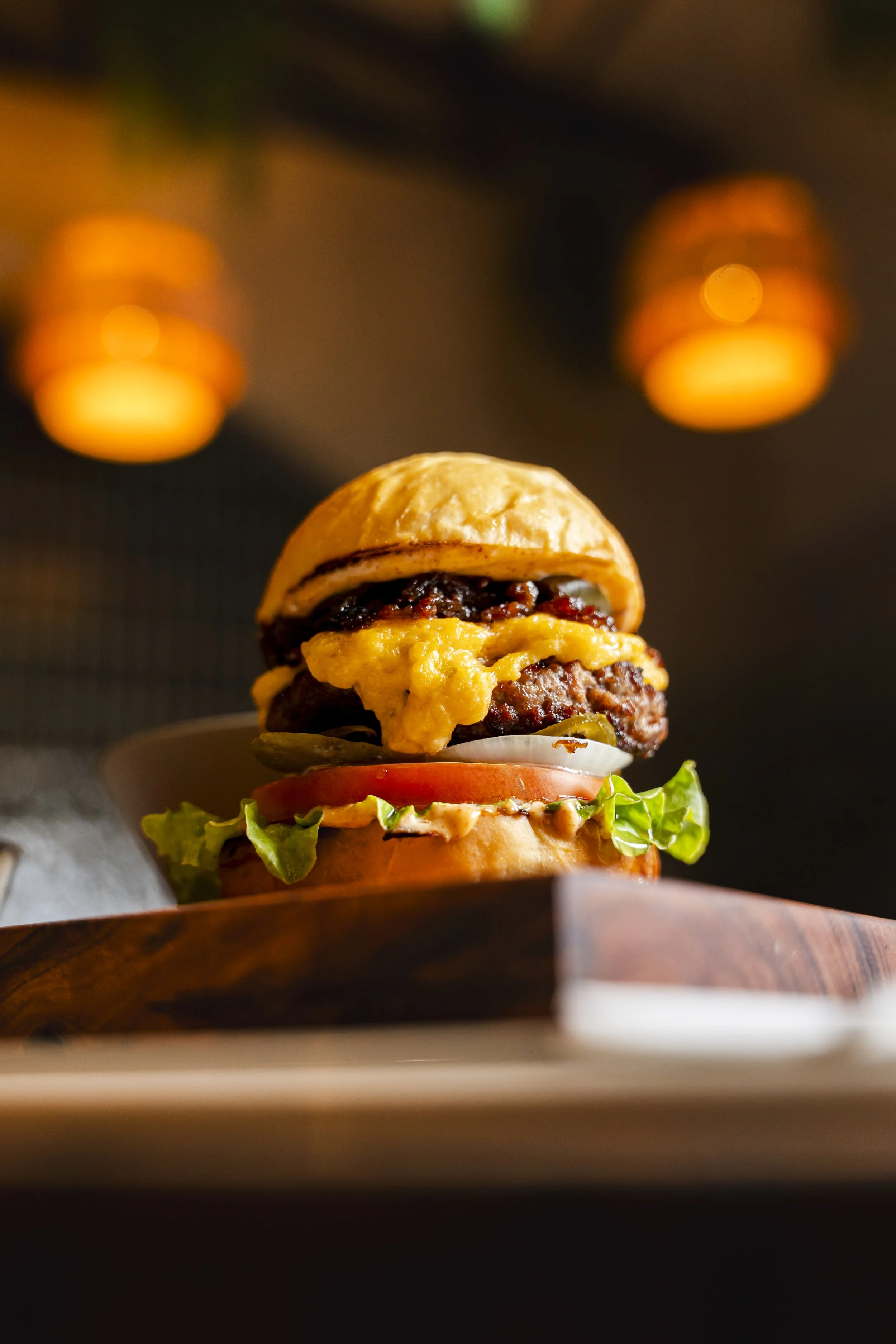 A close-up of a burger with a bun, lettuce, tomato, pickles, a beef patty, cheddar cheese, and caramelized onions, served on a wooden platter inside a restaurant with warm lighting.