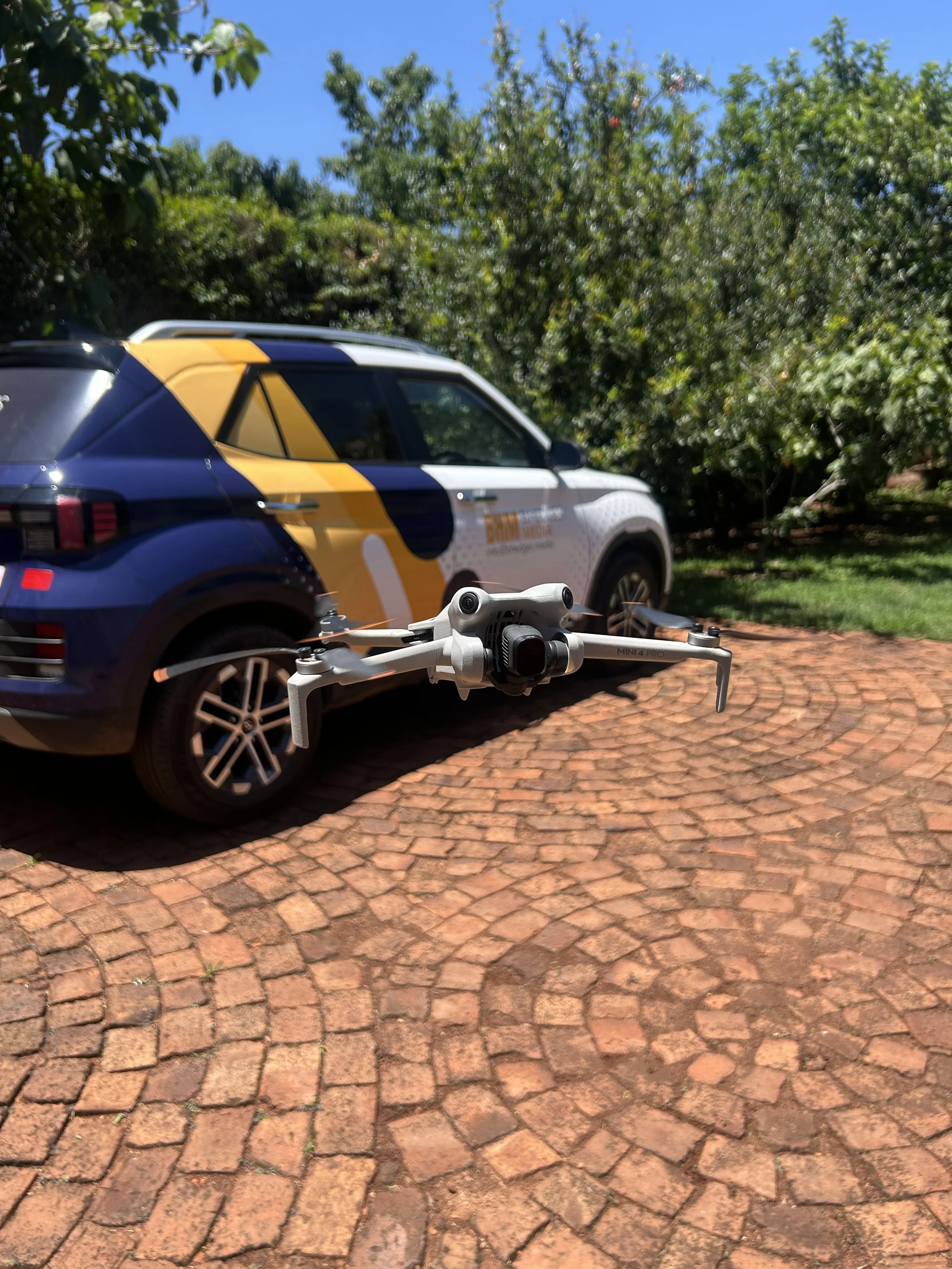 A drone flying in front of a colorful vehicle with a design featuring yellow, white, and black shapes, parked on a brick driveway with green trees in the background.