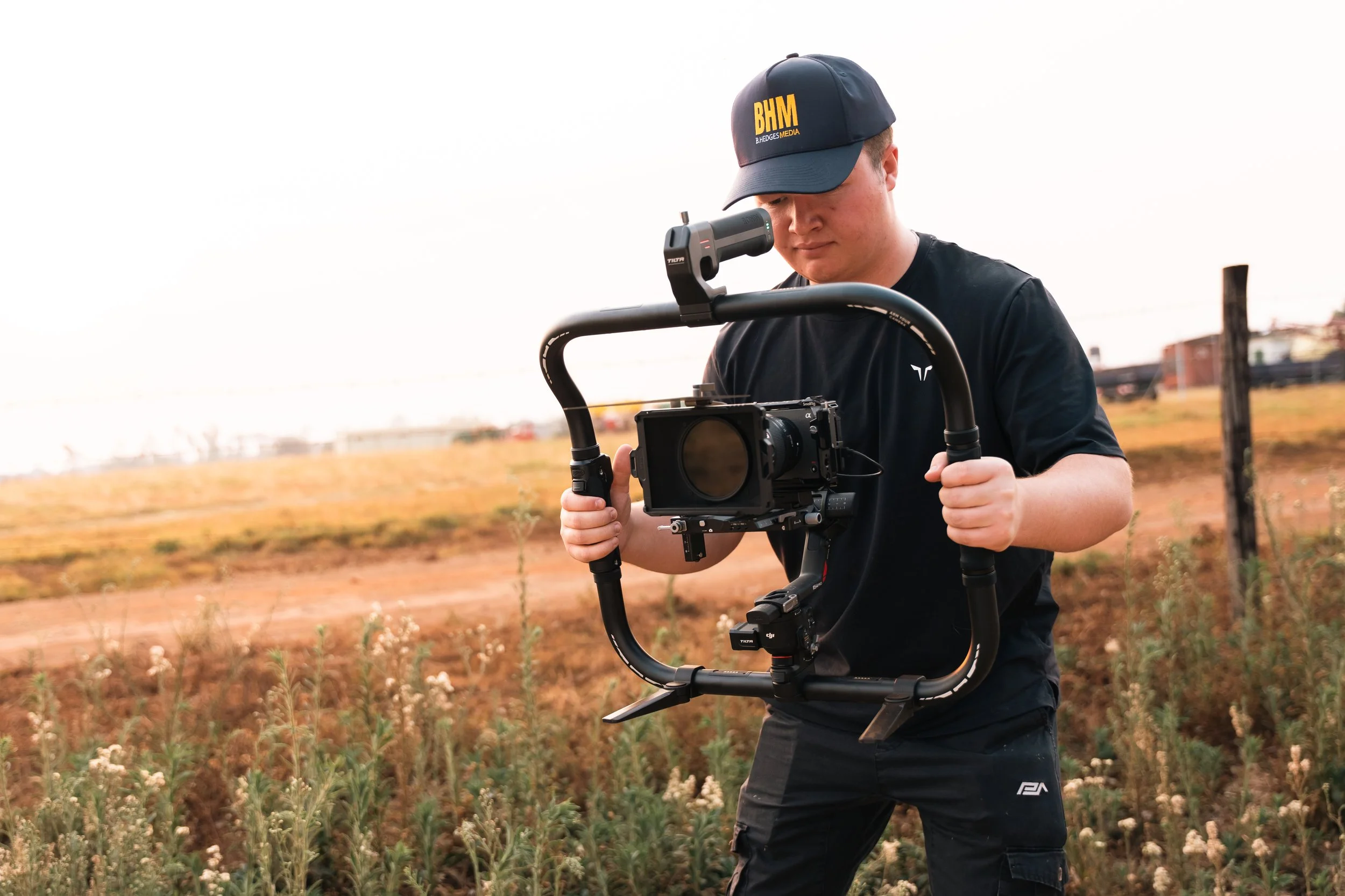 Person operating a camera stabilizer outdoors in a field with dry grass and plants.