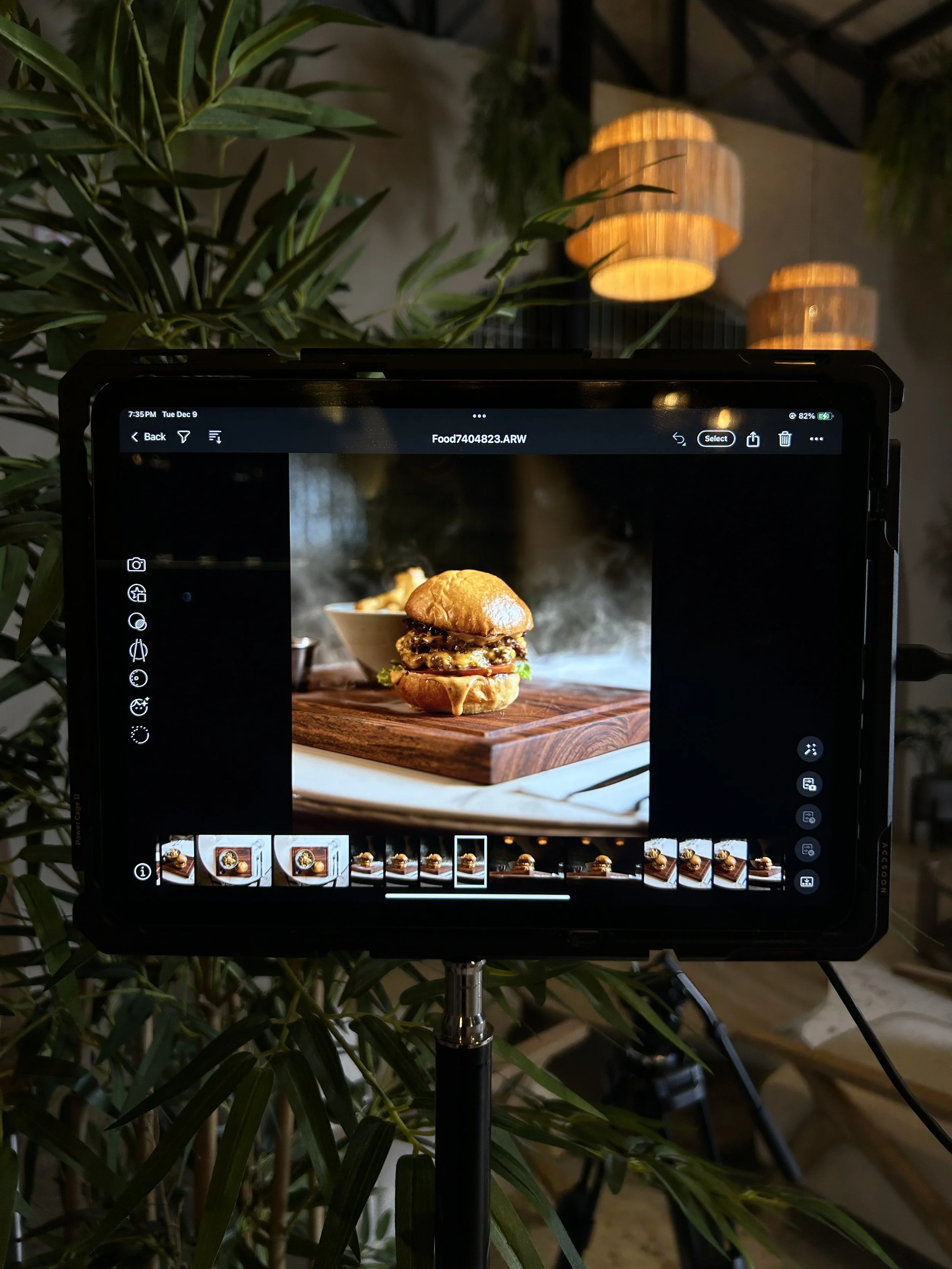 A digital screen displaying a close-up photo of a cheeseburger with lettuce and melted cheese, placed on a wooden cutting board. The background includes plants and hanging lamps.