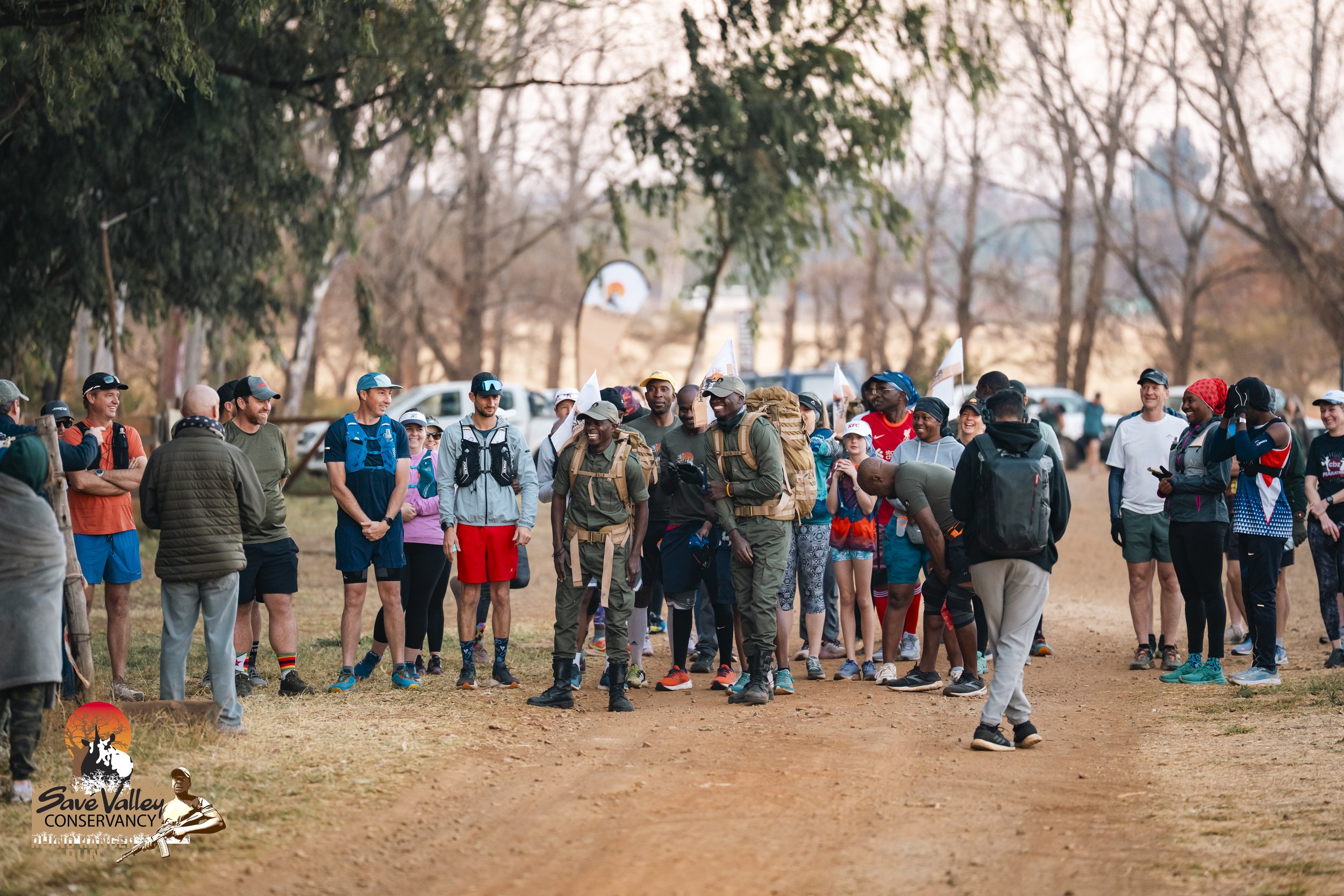 A group of hikers and runners, dressed in athletic gear and backpacks, gathered outdoors on a dirt path surrounded by trees during sunset, participating in the Save Valley Conservancy event.