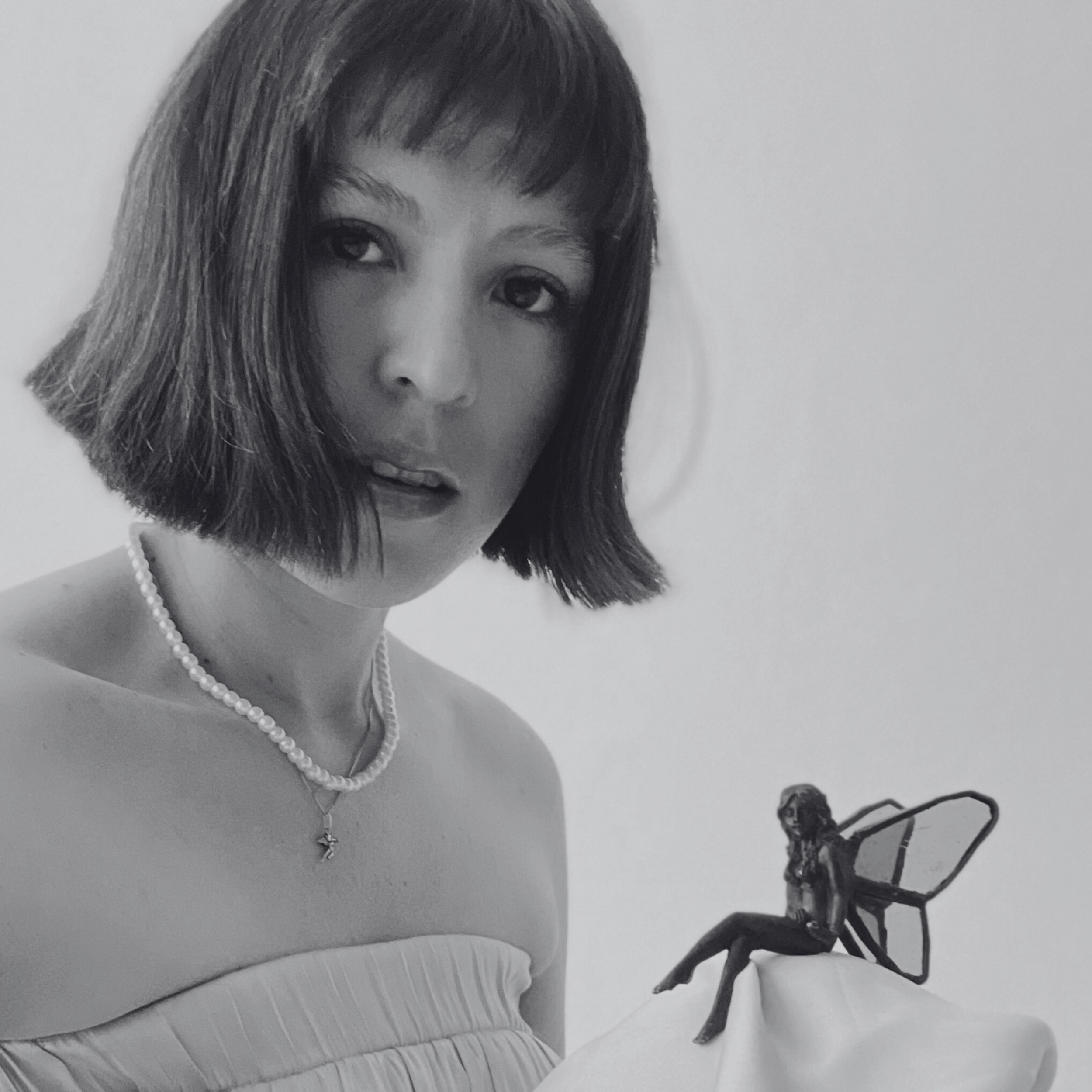 Black and white photograph of a woman with a short bob haircut and pearl necklace, holding a small fairy figurine with wings on her hand.