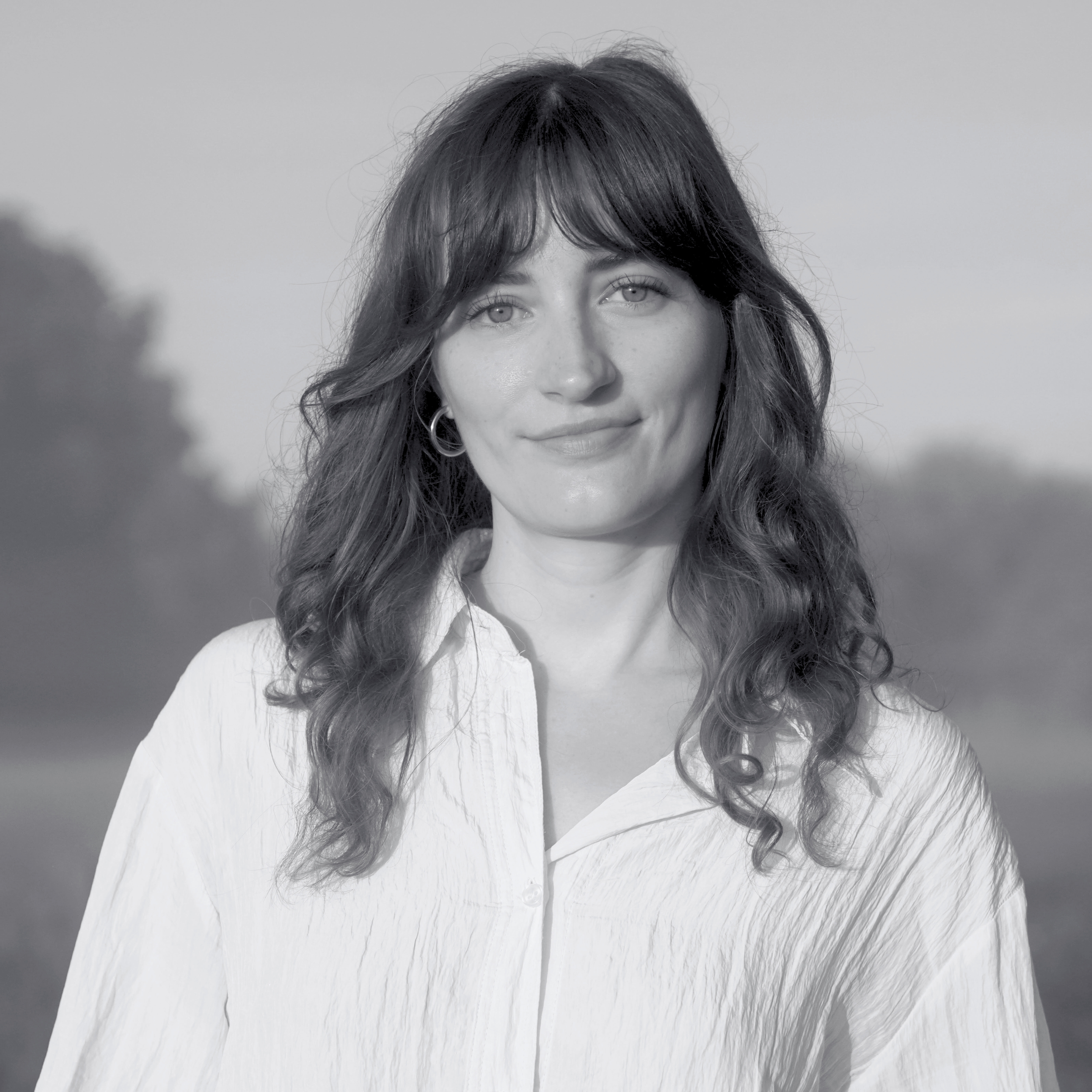 Black and white photo of a woman with long hair and bangs wearing a white shirt, standing outdoors with a blurred background.