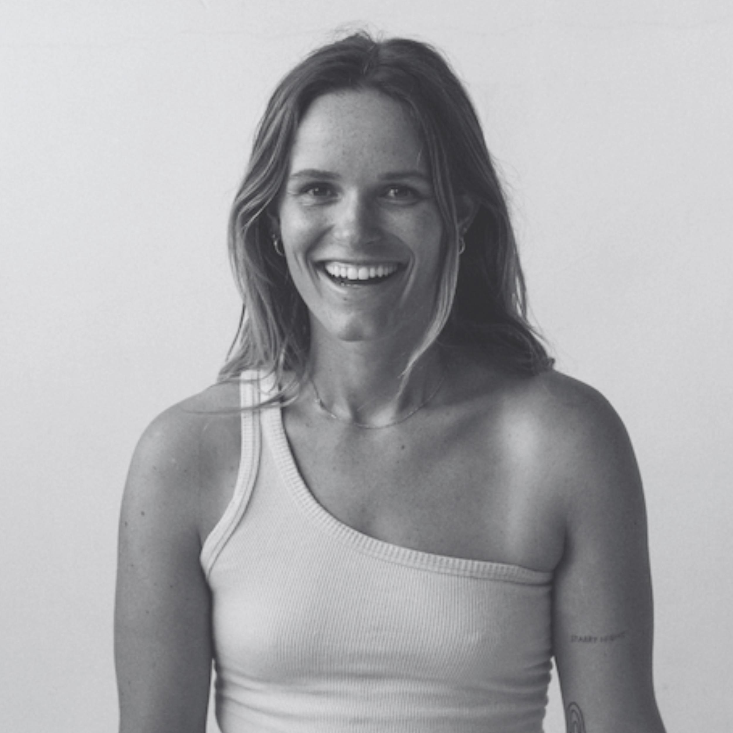 Black and white photo of a smiling woman in a one-shoulder top with a tattoo on her arm, with long hair and simple jewelry.