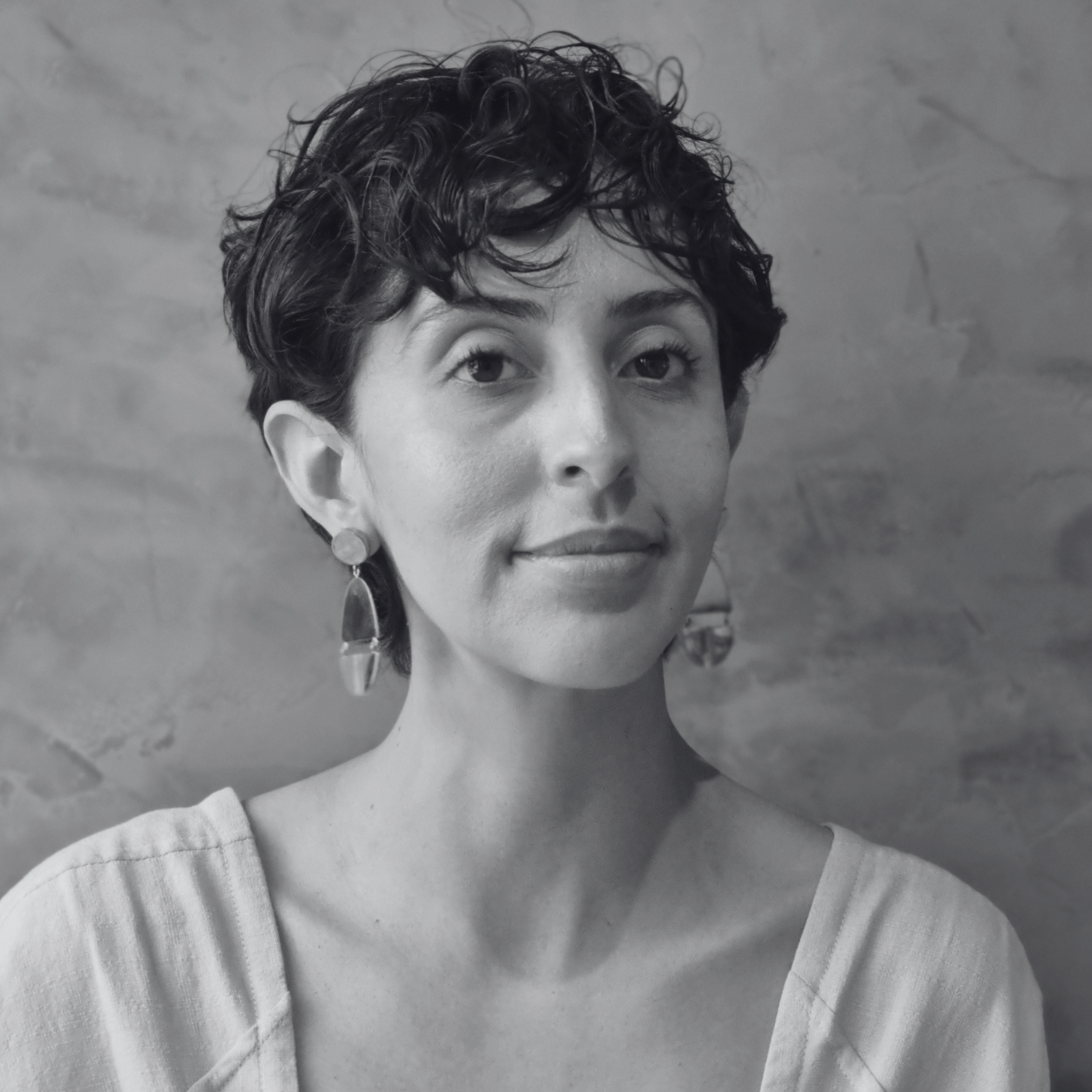 Black and white portrait of a person with short curly hair, wearing earrings and a light-colored top, looking at the camera with a neutral expression.